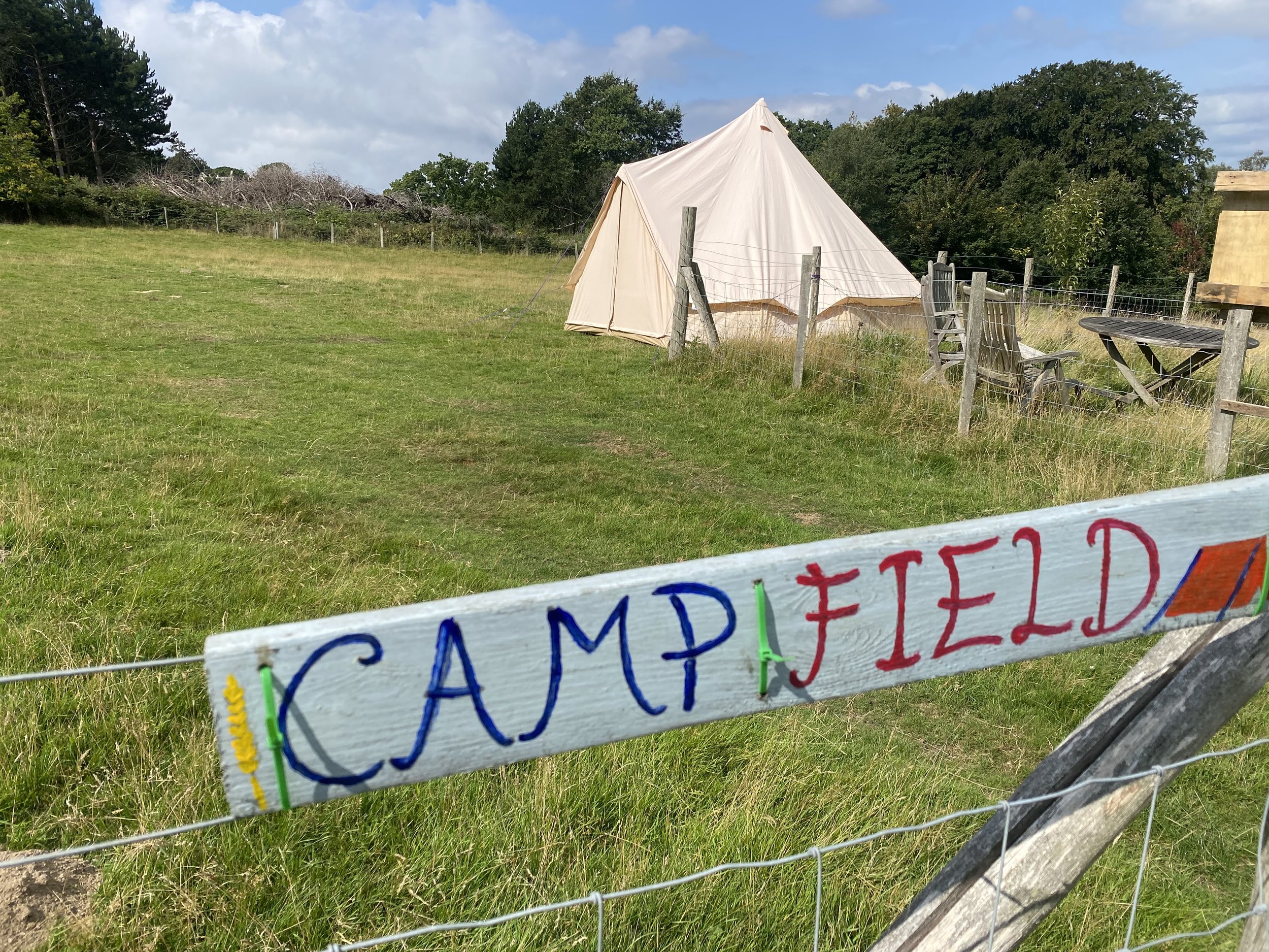 A grassy field with a white tent near a wooden fence. There are outdoor chairs and tables, and a sign that reads 'CAMPFIELD' in colorful letters. Trees and a cloudy sky are in the background.