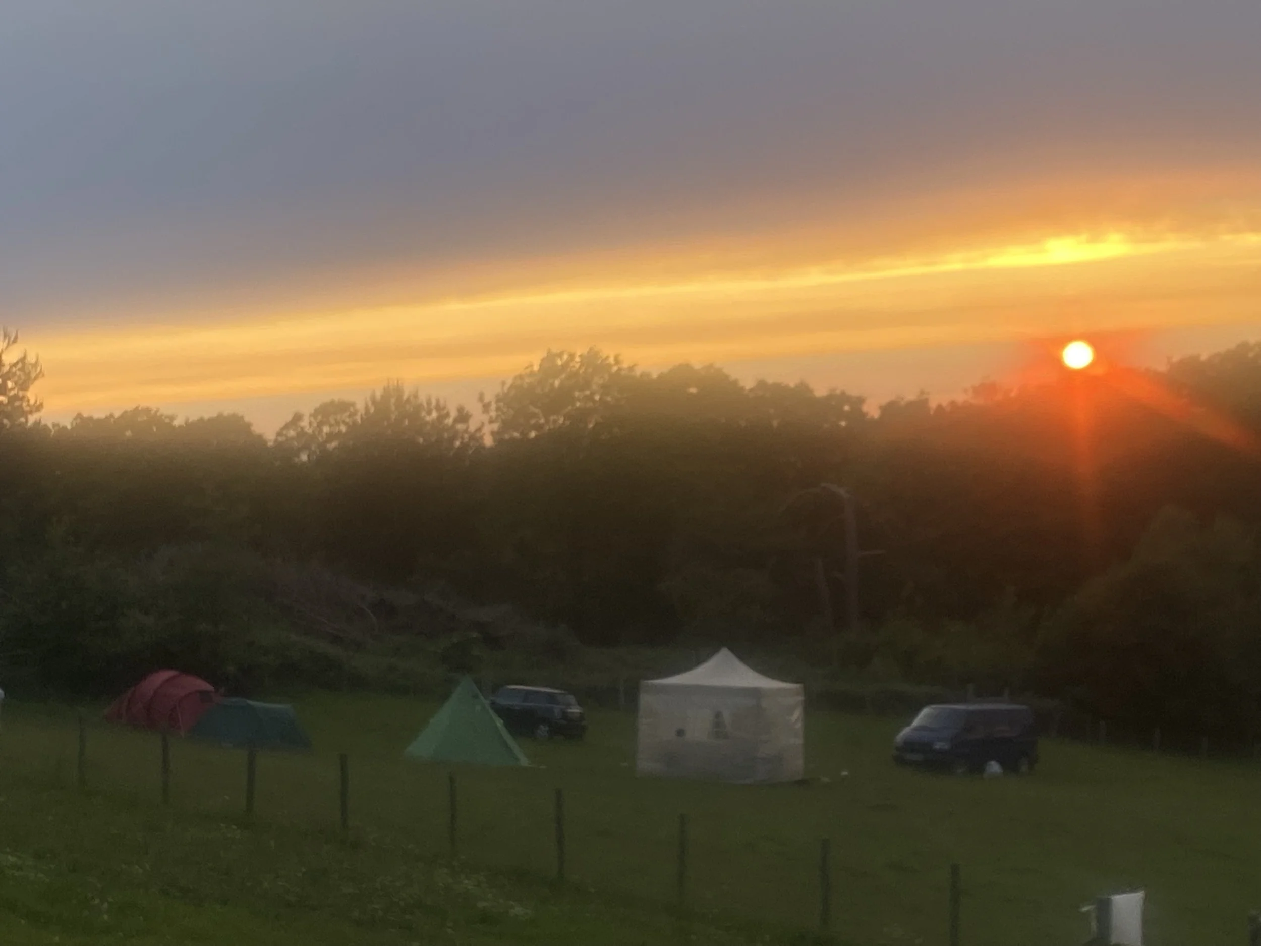 Sunset over a grassy area with tents, vehicles, a white canopy, and trees in the background.