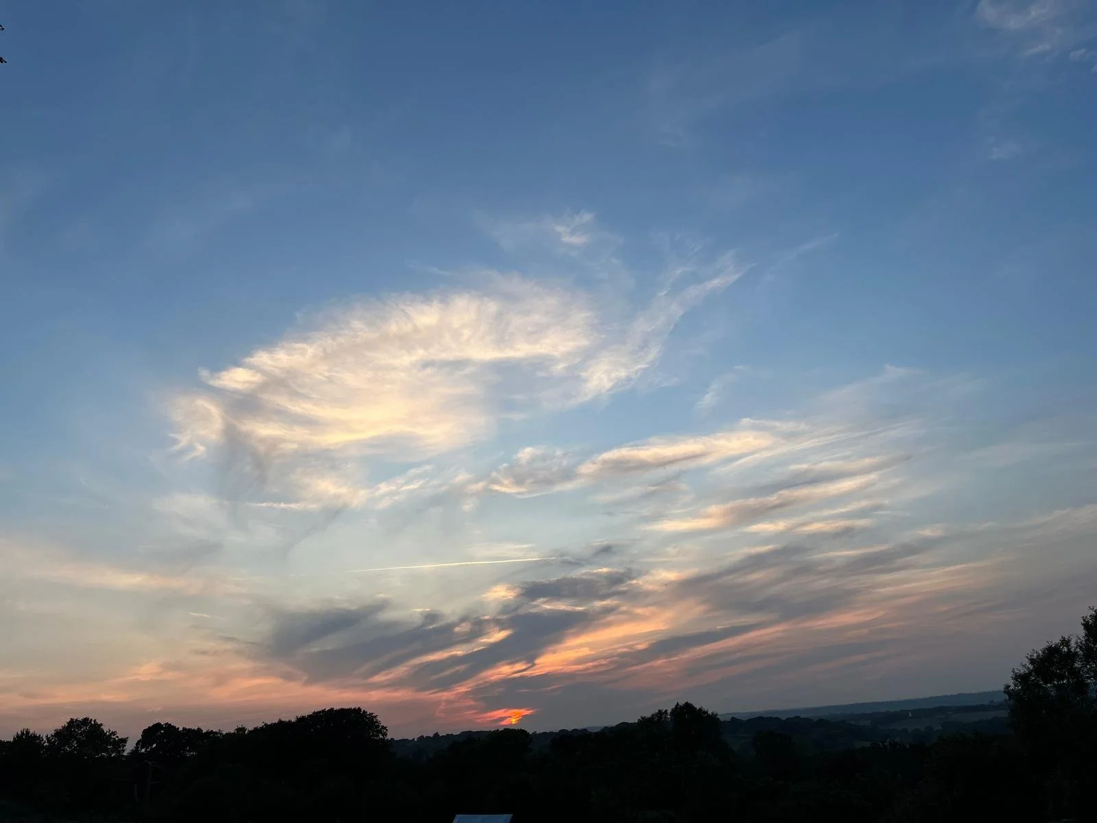 A scenic view of the sky during sunset with soft clouds and a faint orange glow near the horizon, with silhouettes of trees and a landscape in the foreground.
