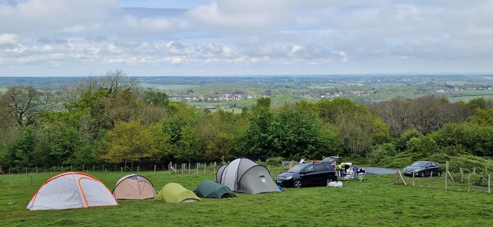 Camping scene with tents set up on grassy field, surrounded by trees, and a distant view of a countryside with houses and fields under a partly cloudy sky.