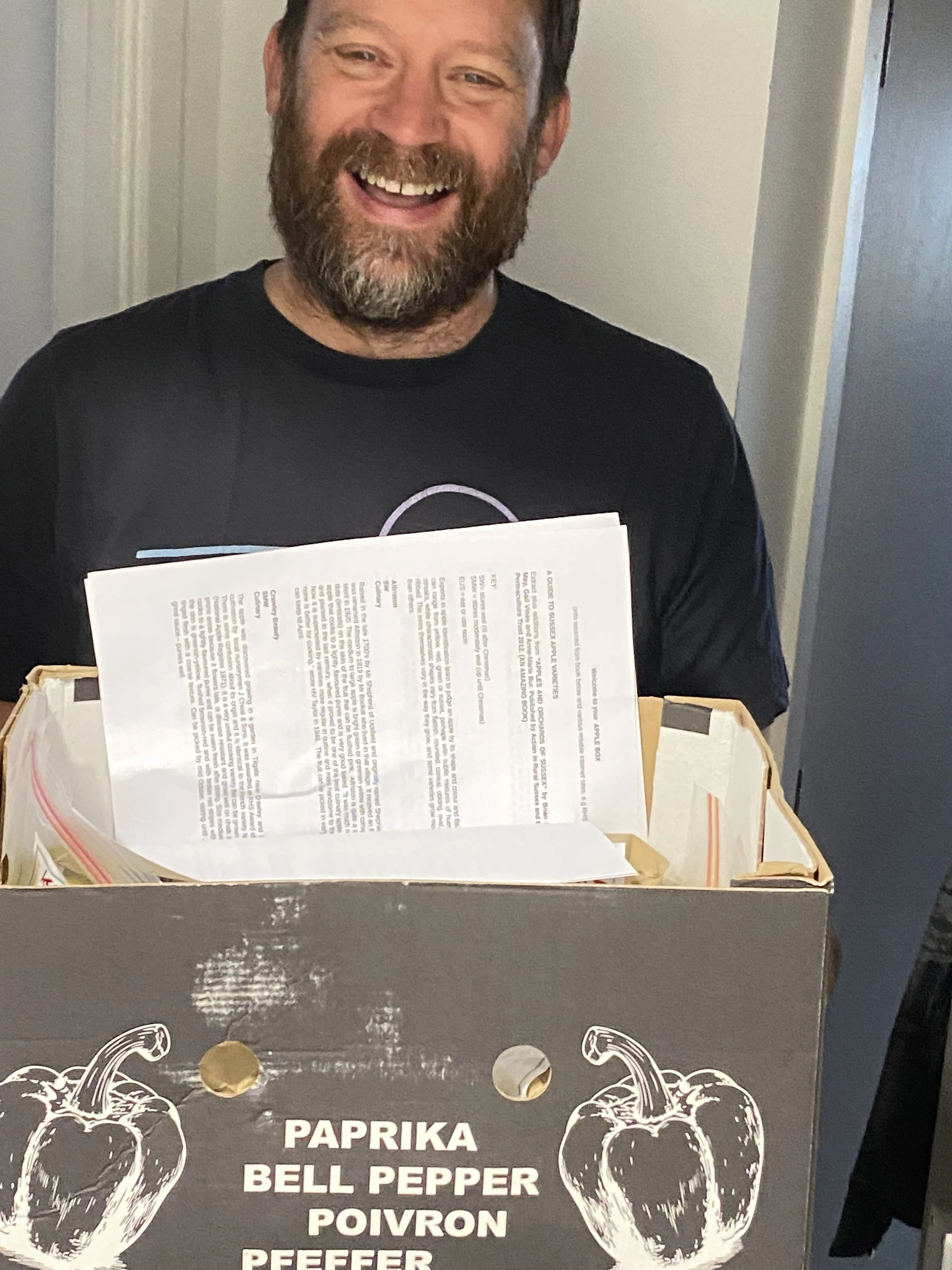 A man with a beard smiling while holding a box filled with papers. The box has an illustration of a bell pepper and the words 'Paprika, Bell Pepper, Poivron, Peffer' on it.