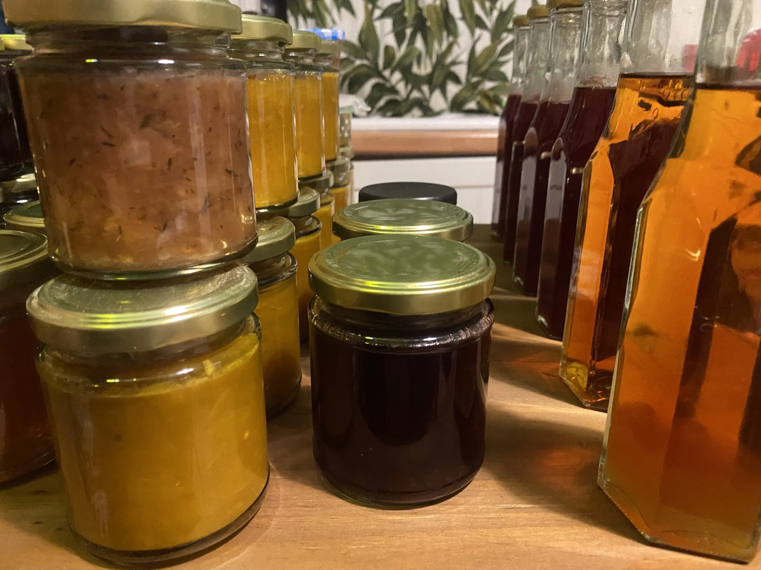 A collection of glass jars and bottles filled with various jams, preserves, and liquids, arranged on a wooden surface in a kitchen.