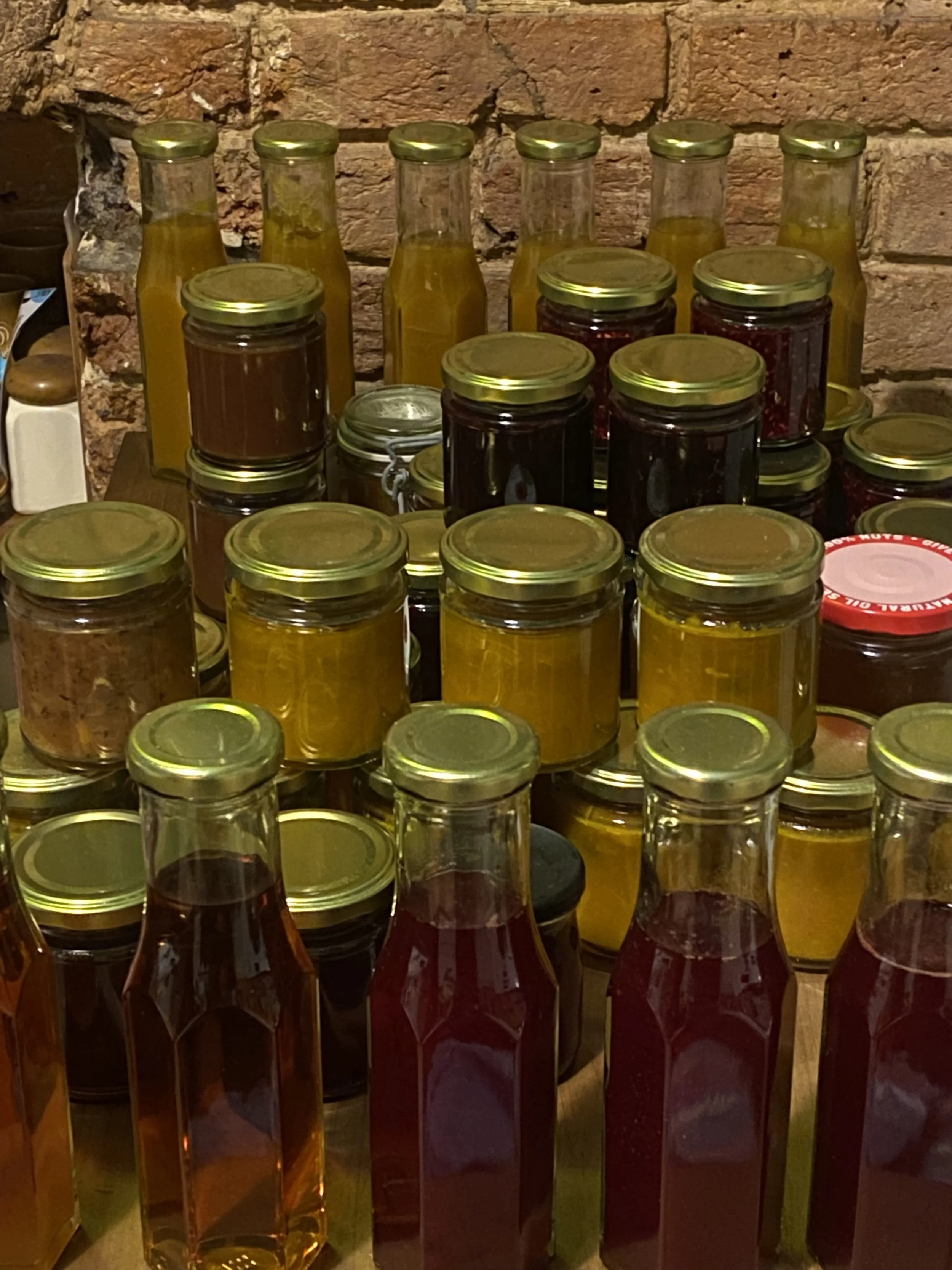 A variety of glass jars filled with colorful preserves, jams, or sauces, arranged on a wooden surface against a brick wall background.
