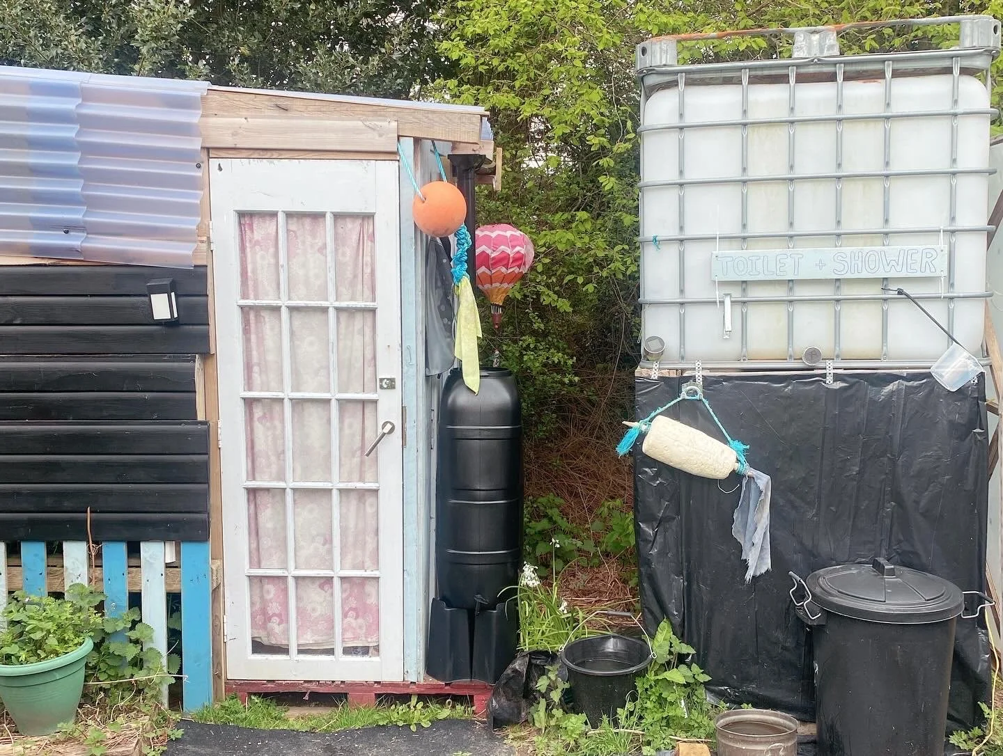 Backyard shed with a sign indicating 'Toilet + Shower,' black water tanks, watering cans, and greenery.