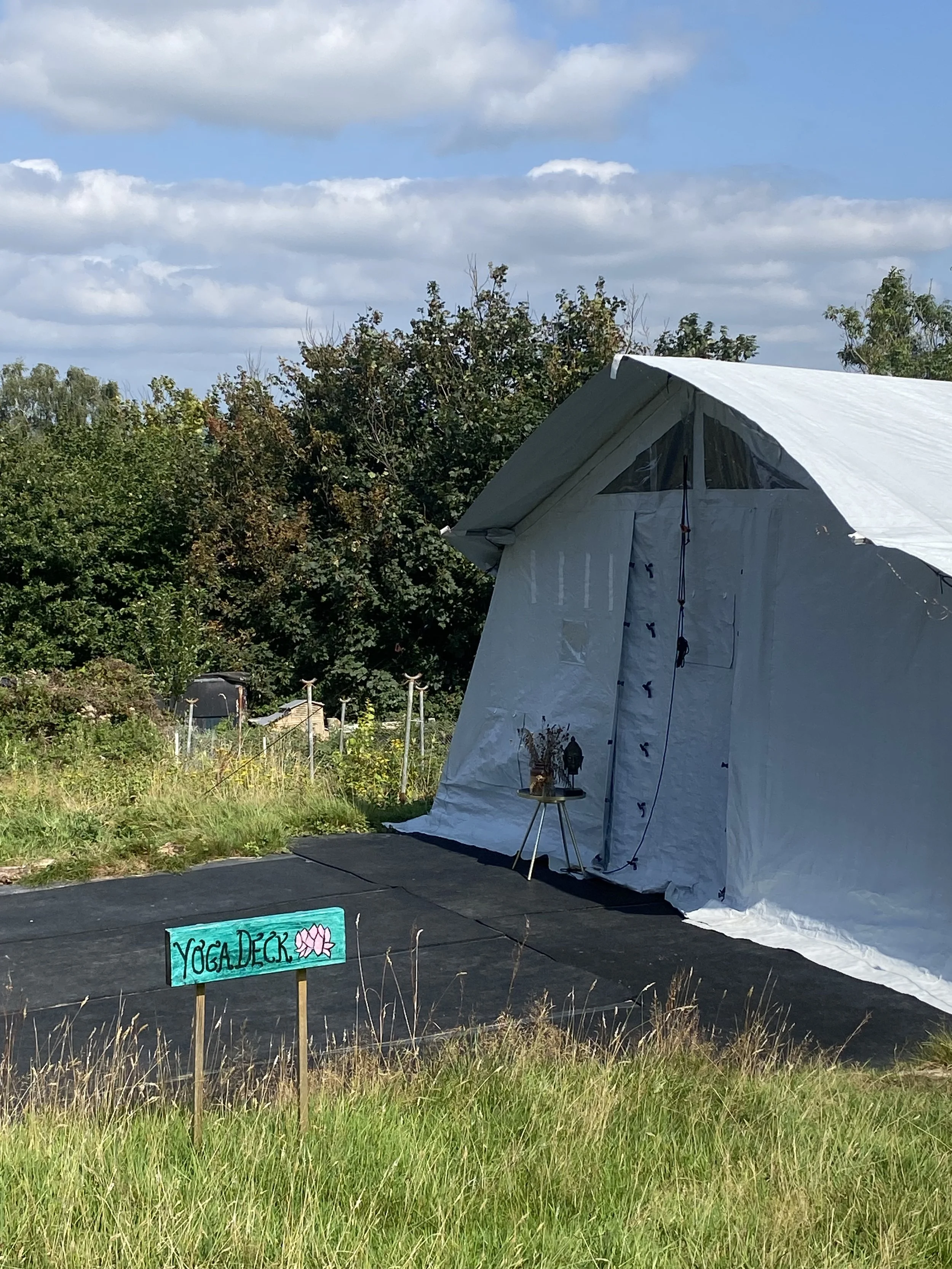 An outdoor yoga deck with a black mat surface set up on a grassy field next to a large white tent with a sign reading 'YOGA DECK' and a pink lotus flower illustration, with trees and a partly cloudy sky in the background.