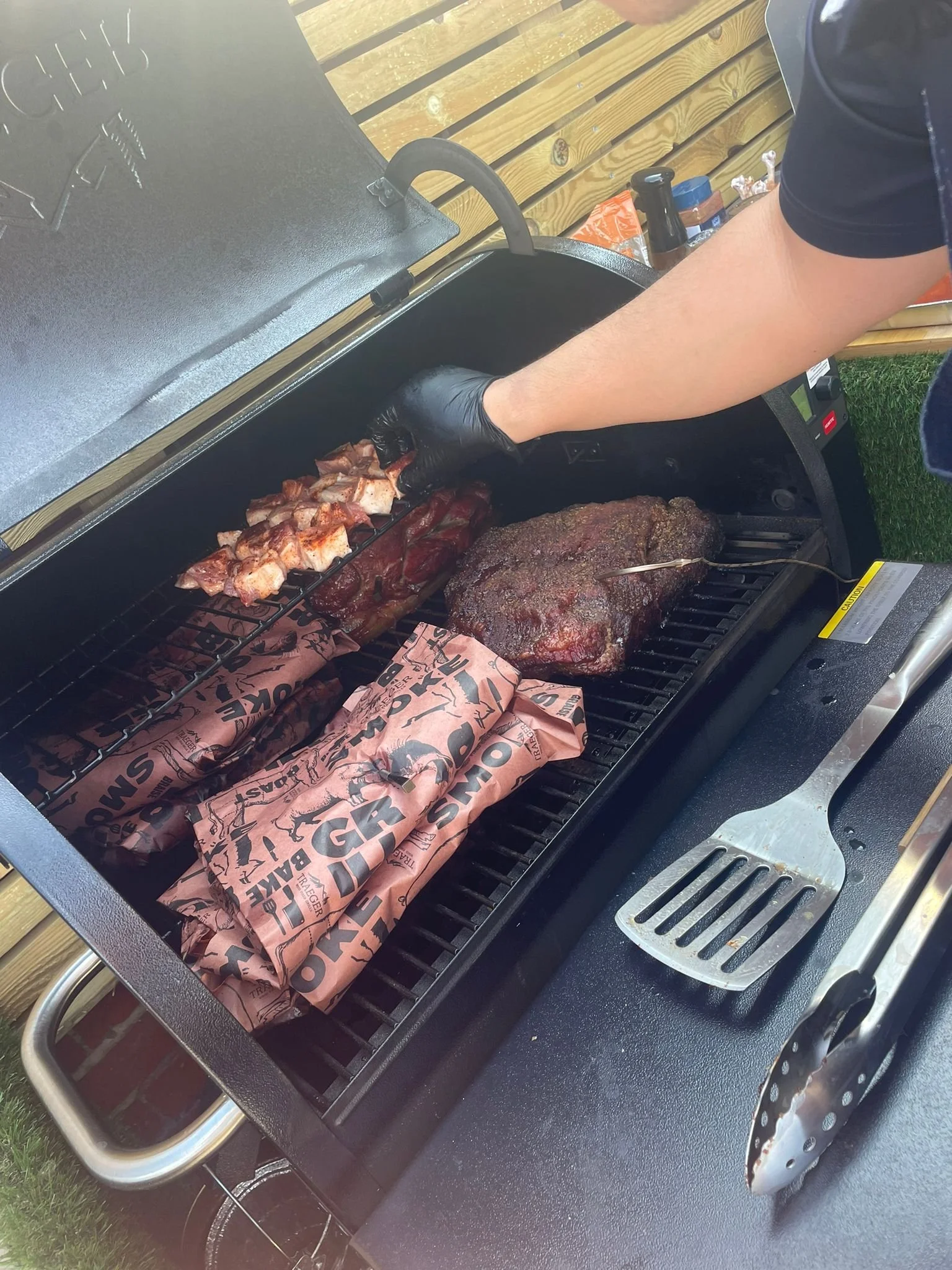 A person wearing black gloves is grilling meat on a barbecue grill, with skewered chicken, a large beef roast, and wrapped packages on the grill. A spatula and tongs are on the side of the grill.