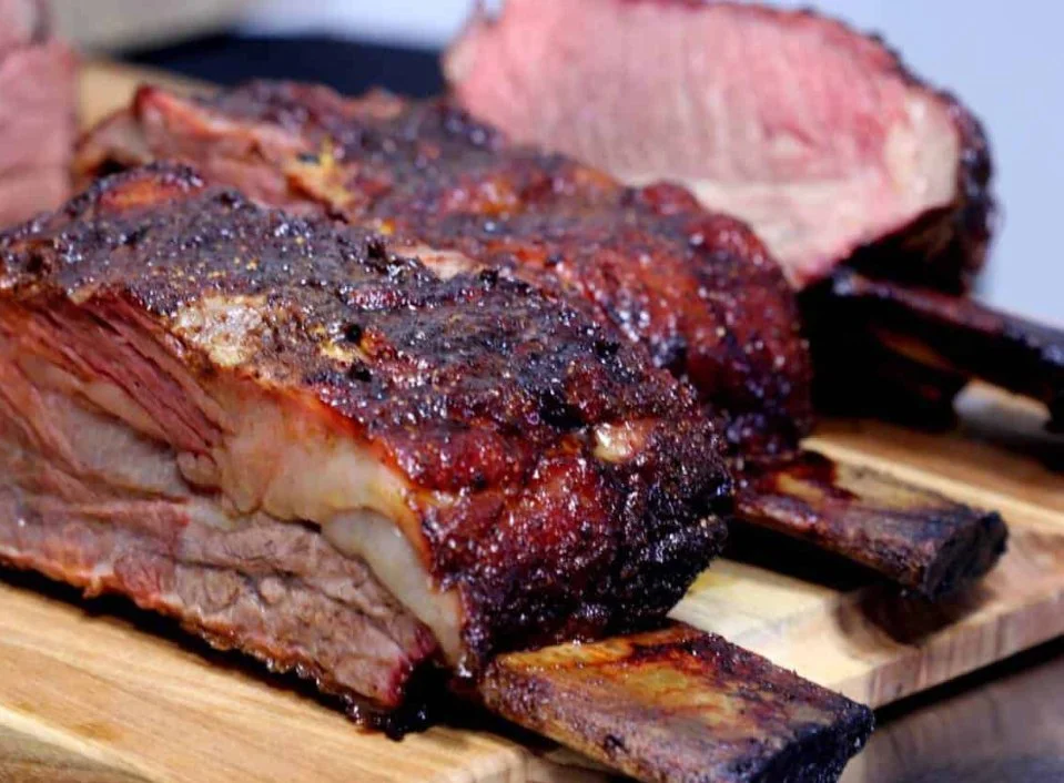 A close-up of cooked beef ribs on a wooden cutting board, with some meat sliced to show the interior.