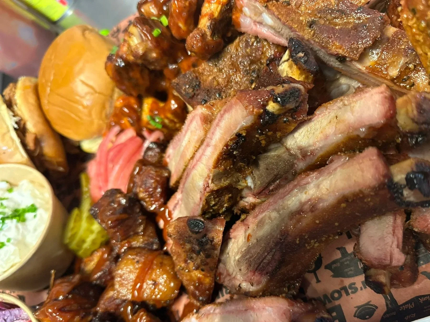 Close-up of barbecued meats including ribs, chicken, and brisket, with side dishes like potato salad and a pickle.