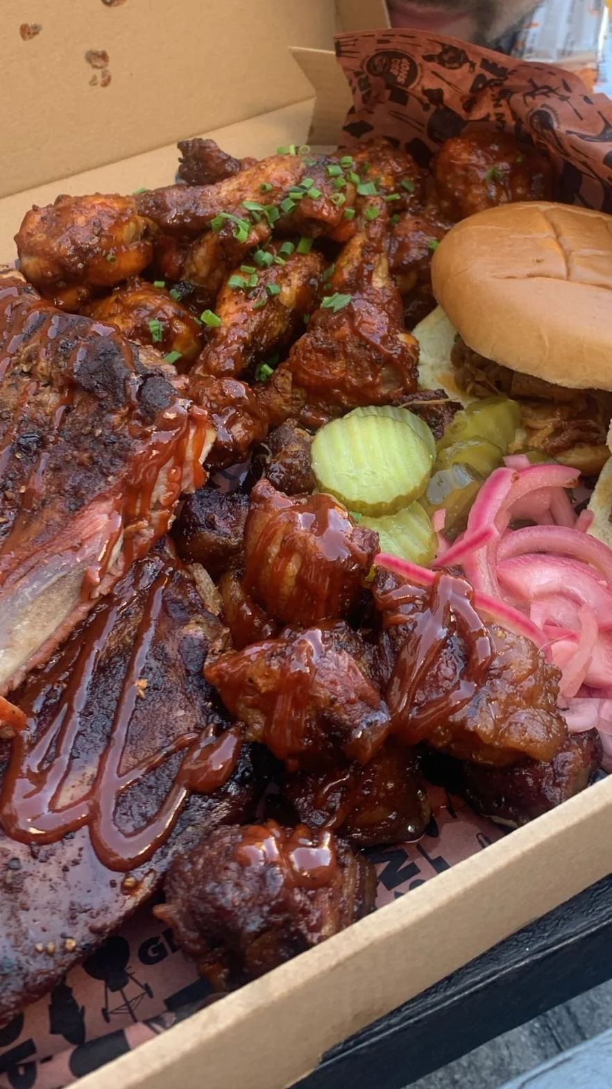 Barbecue platter with glazed ribs, fried chicken wings topped with chopped green onions, pickles, sliced onions, and a small burger with a bun and beef patty, served in a cardboard takeout box.