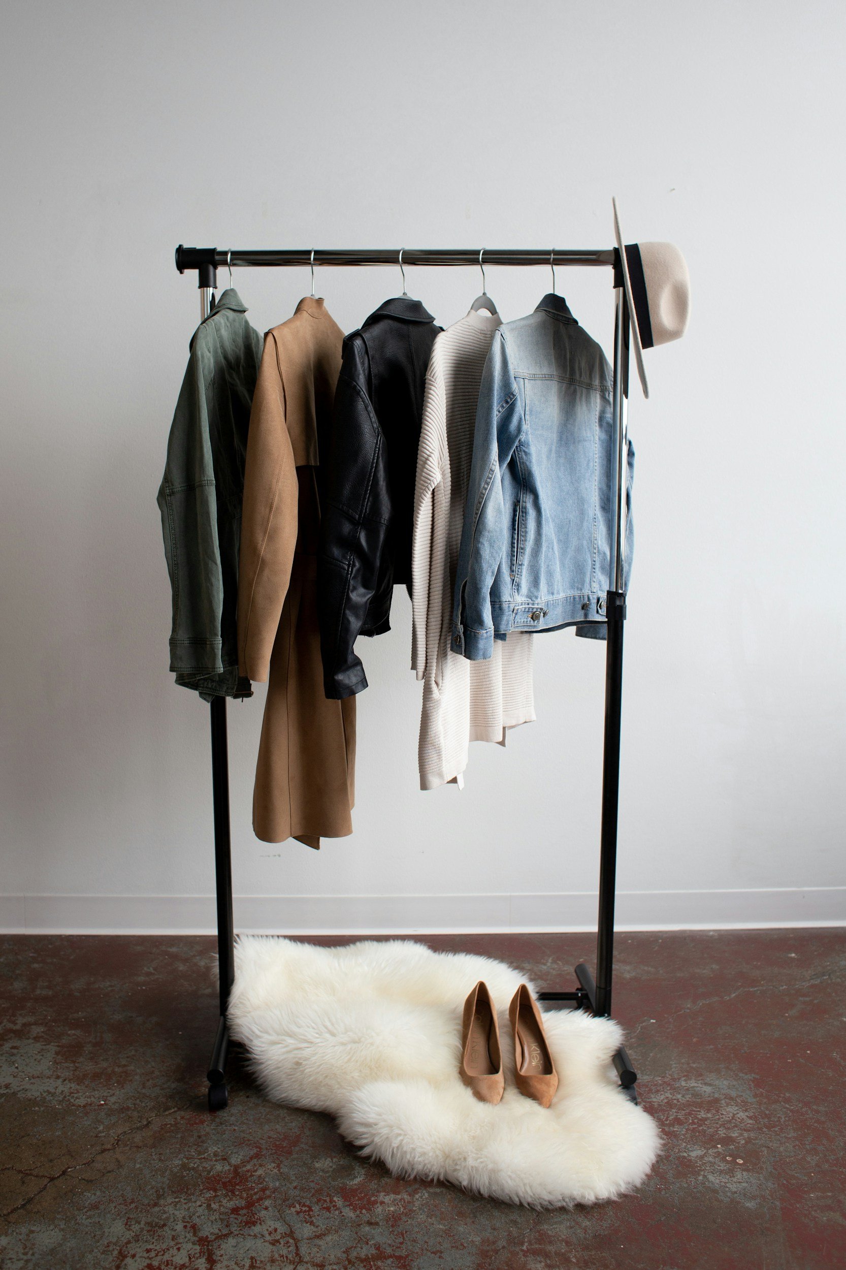 Clothing rack with five jackets and sweaters, a beige hat hanging on the right side, a fluffy white rug with beige high heels underneath, all against a plain white wall and a concrete floor.