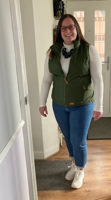 Woman with glasses smiling, wearing a white long sleeve shirt, green vest, blue jeans, white sneakers, and a black necklace, standing inside a house.