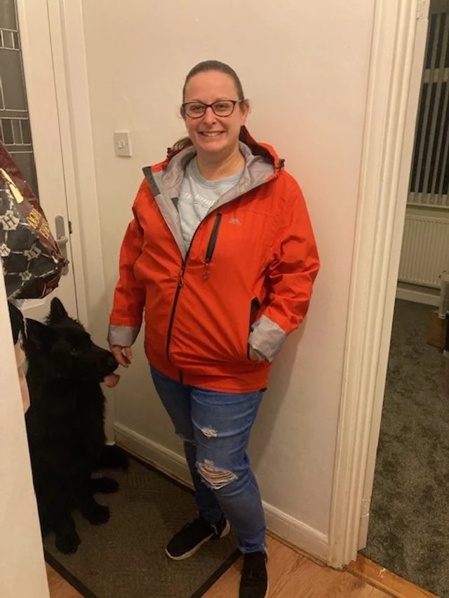 A woman with glasses and a ponytail smiling in a doorway, wearing an orange jacket and ripped jeans, holding a black dog on a leash.