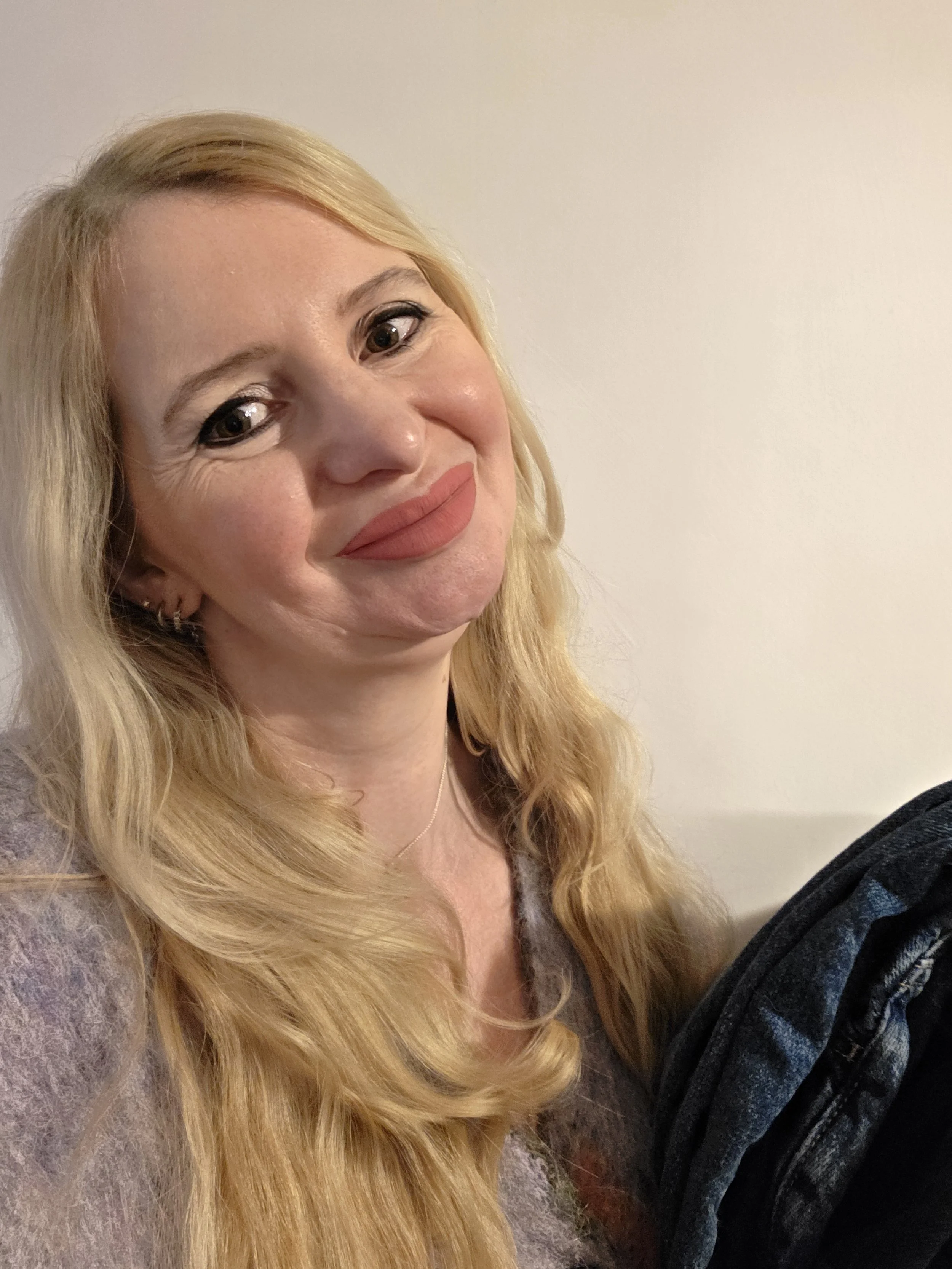 Close-up of a woman with long blonde hair smiling softly, wearing makeup with nude lipstick, earrings, and a delicate necklace, against a plain light-colored wall.