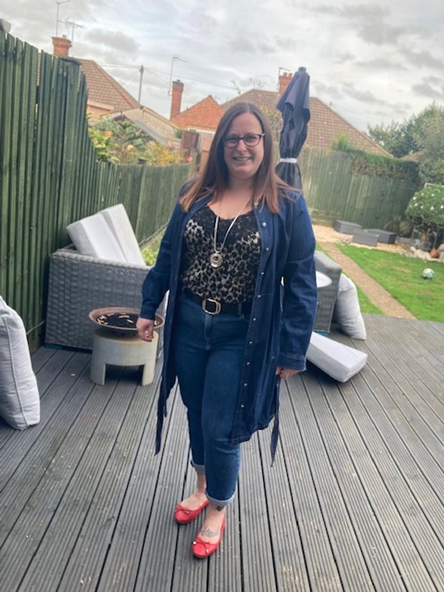 A woman standing on a wooden deck in a backyard, wearing red shoes, a leopard print top, jeans, and a blue jacket, smiling at the camera.