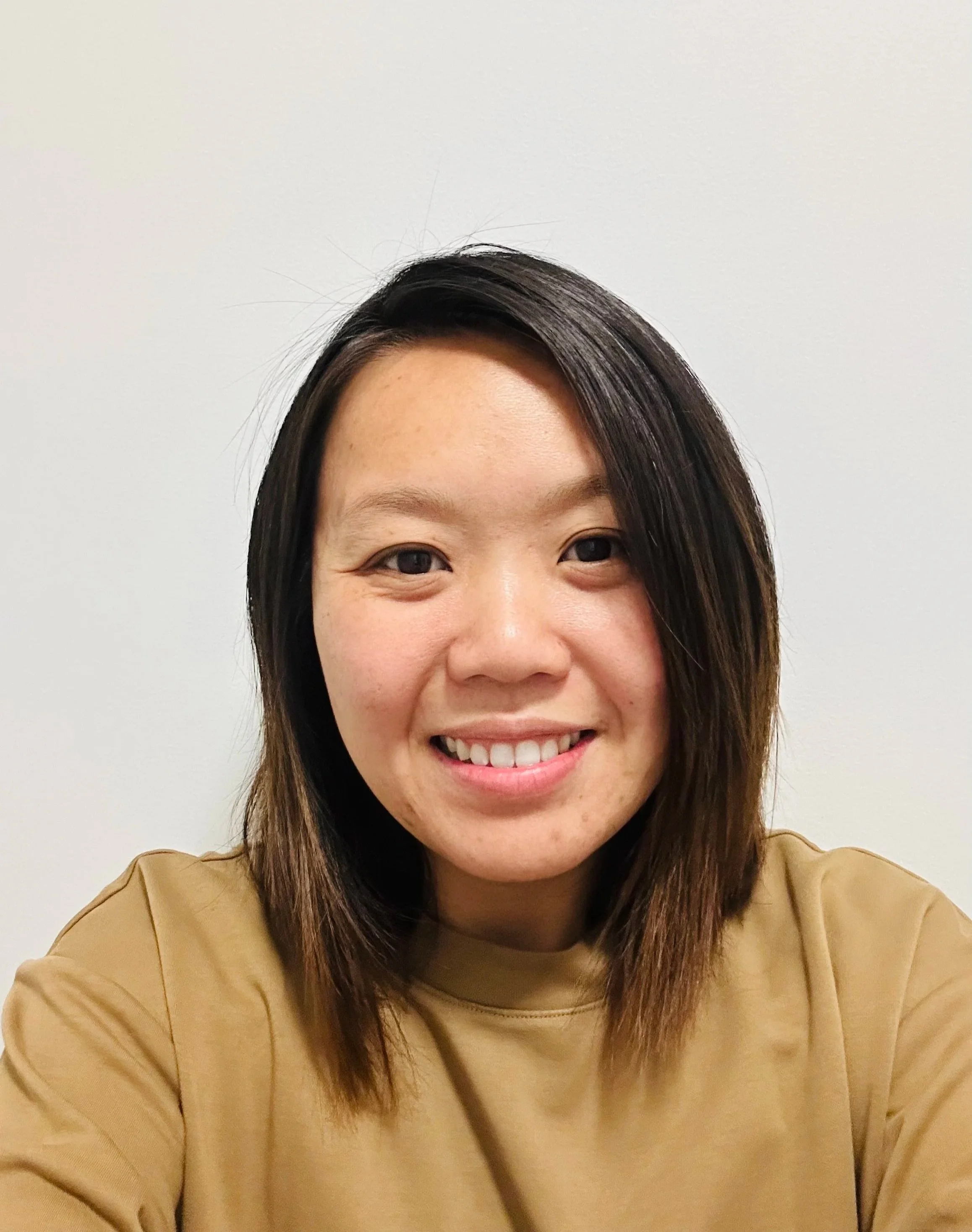 A woman smiling, wearing a tan top, with shoulder-length dark hair, in front of a plain white wall.