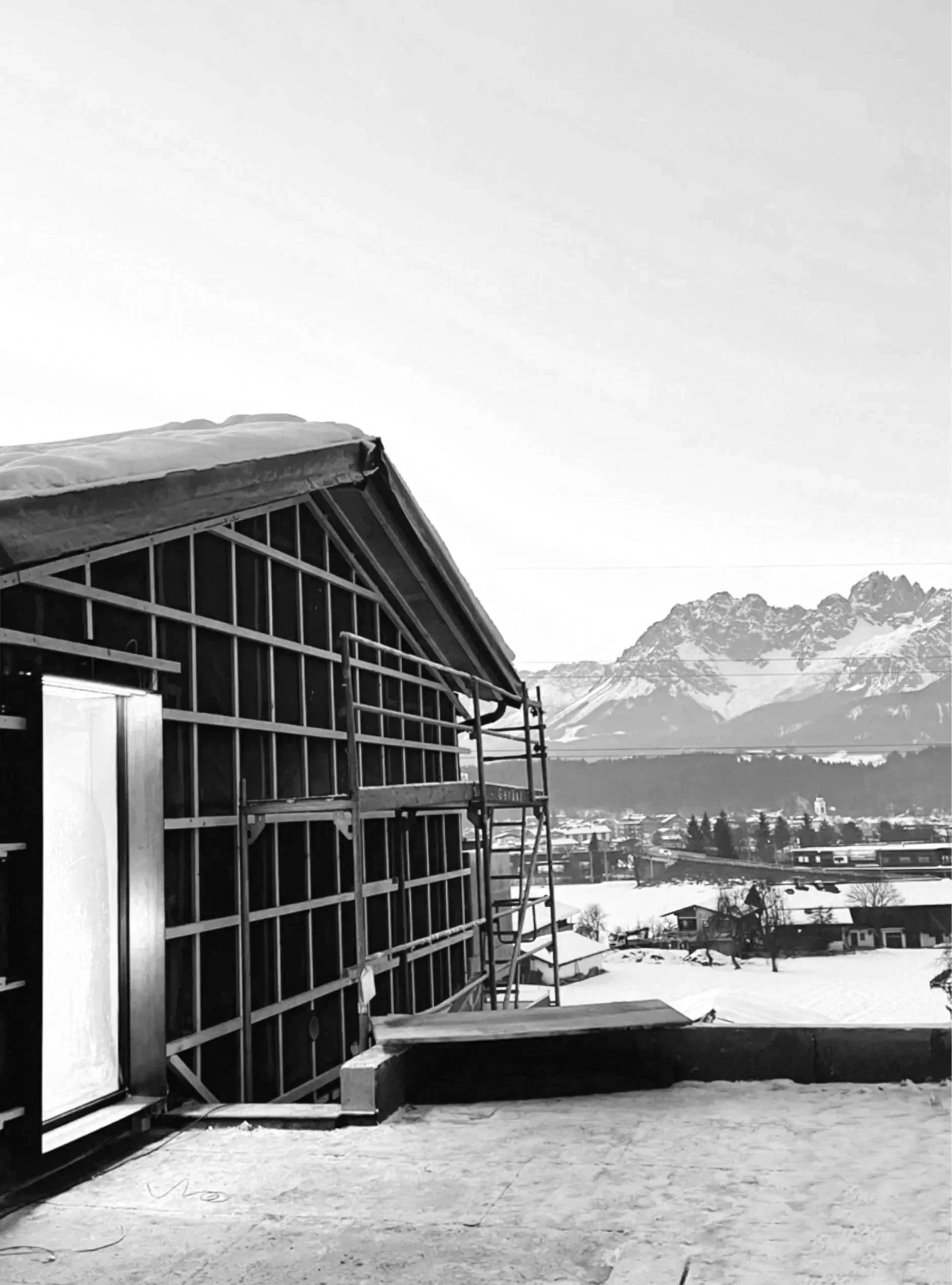 Ein Baugerüst vor einem modernen Gebäude mit einer Berglandschaft im Hintergrund, Schneeflächen auf dem Boden.