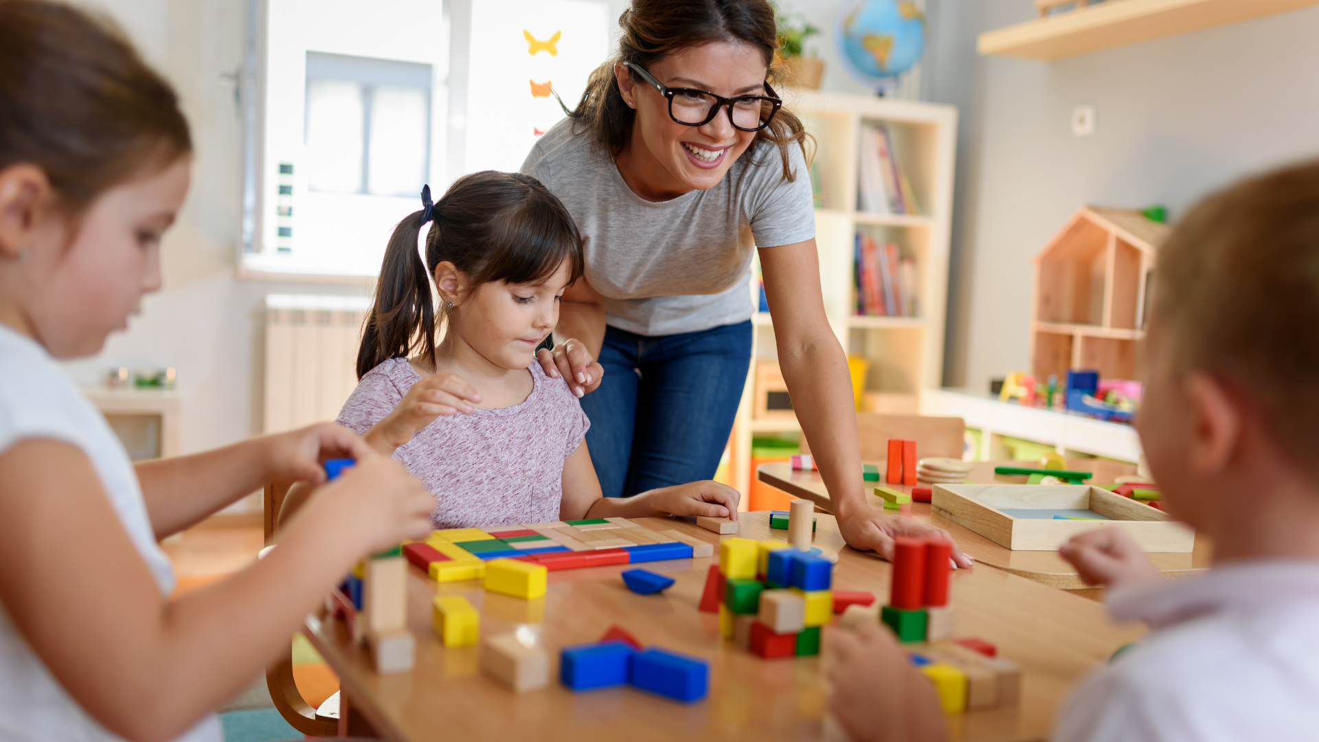 A teacher smiles as she observes three young children playing with colorful wooden building blocks at a table in a classroom.