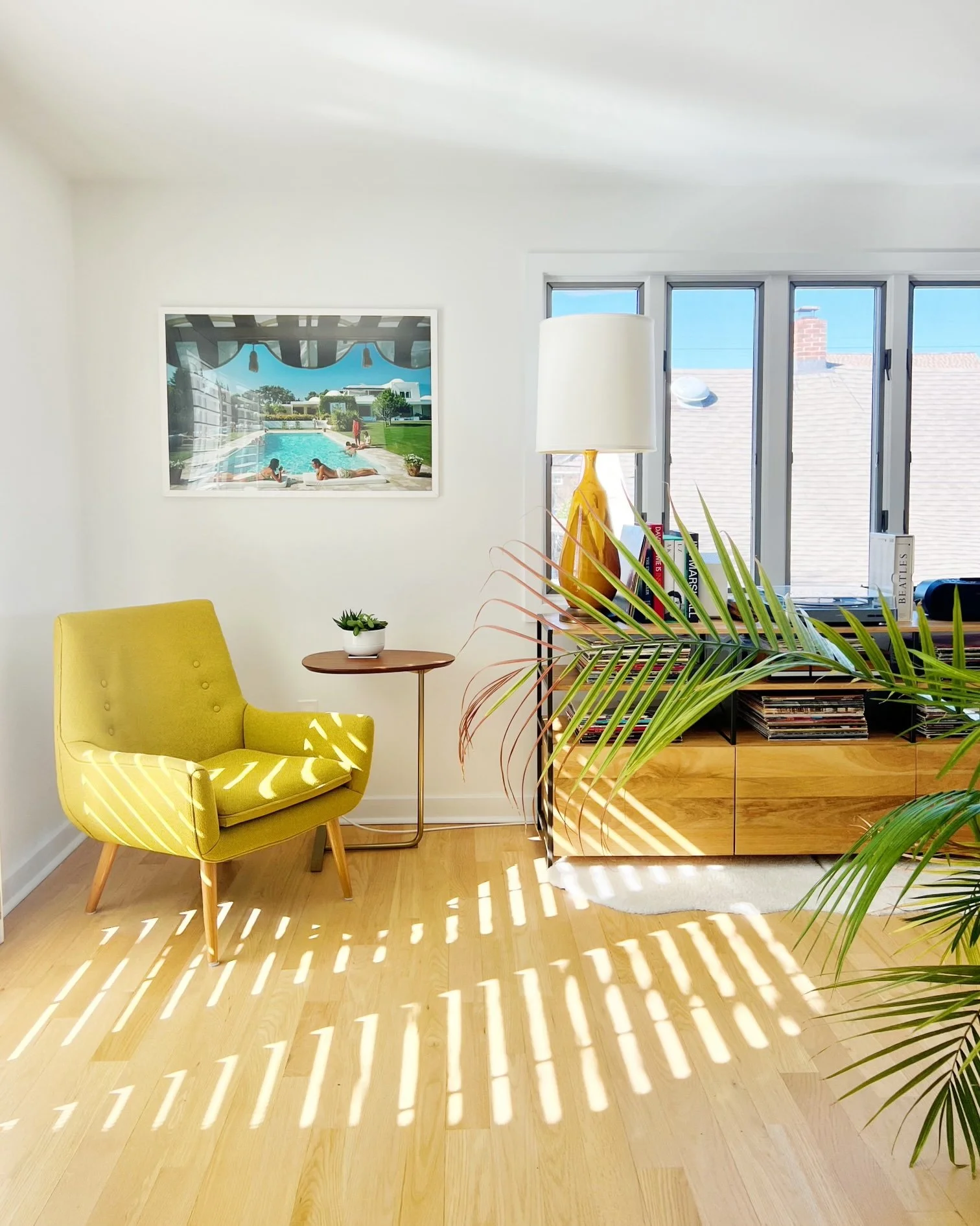 Sunlit living room with yellow armchair, wooden sideboard, large window, potted plant, framed photo of pool scene on wall, lamp on sideboard, and books.