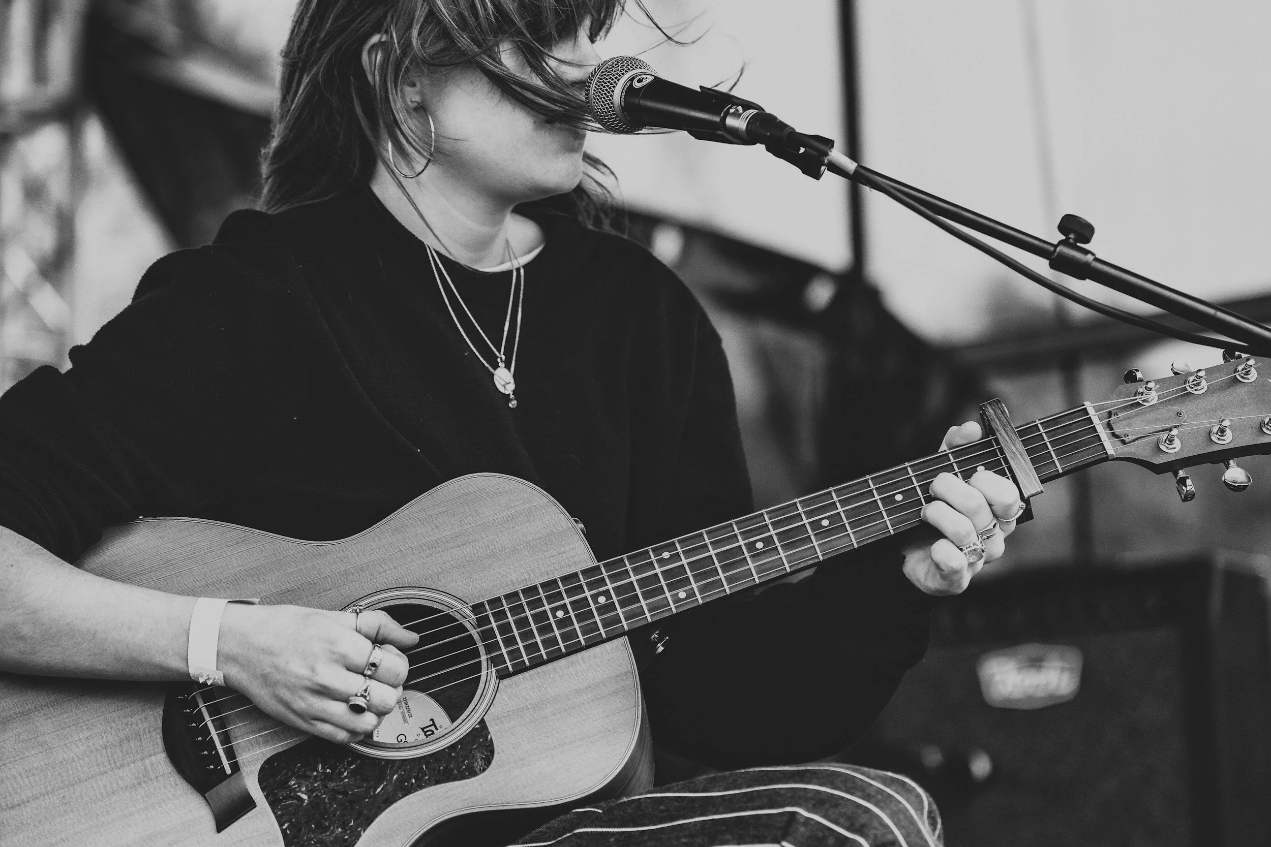 A woman playing an acoustic guitar and singing into a microphone, wearing jewelry and a dark long-sleeve shirt, in a black and white setting.
