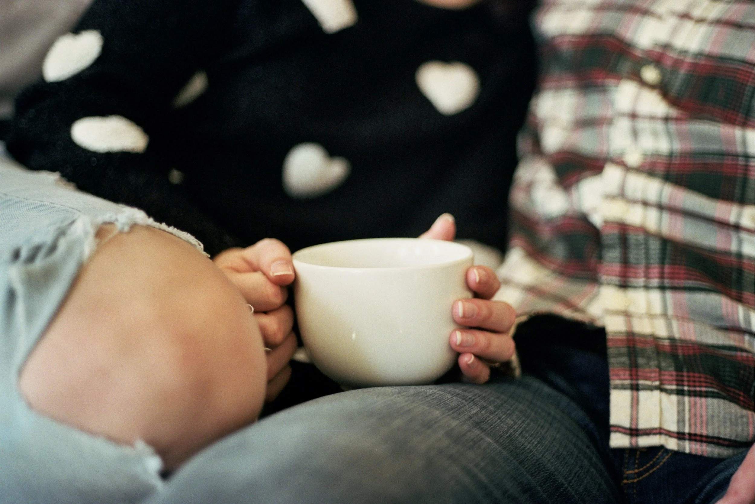 Person holding a white coffee mug, sitting closely with another person wearing a plaid shirt.