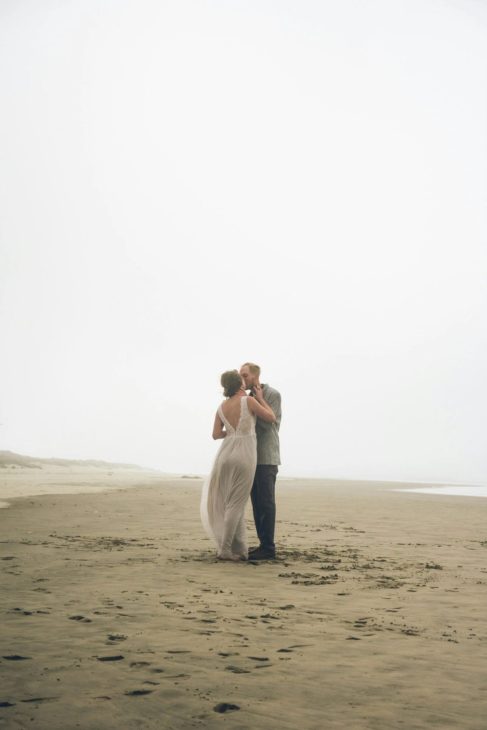 A couple in wedding attire sharing a kiss on a beach with an overcast sky.