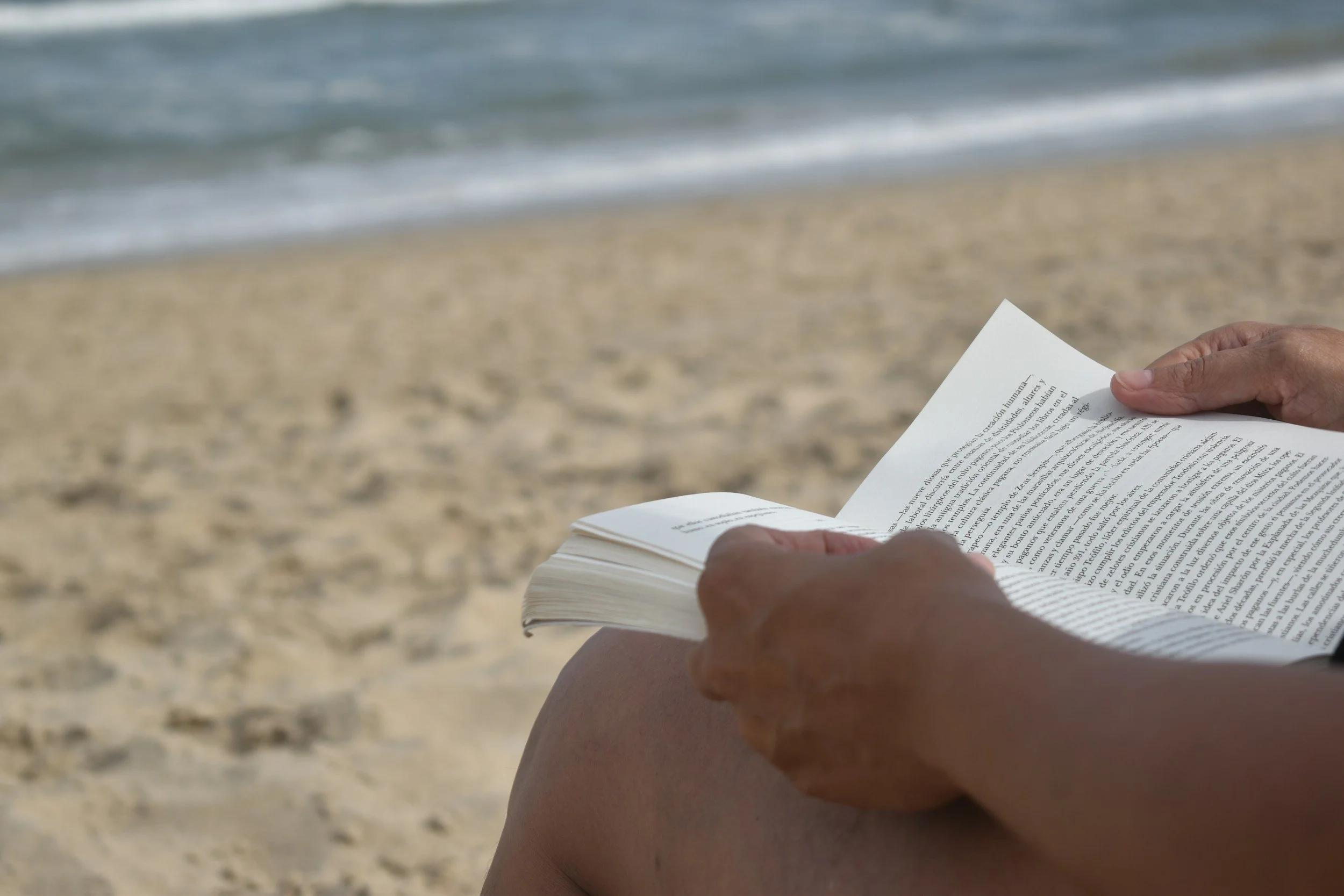 Person reading a book on the beach with sand and ocean in the background.