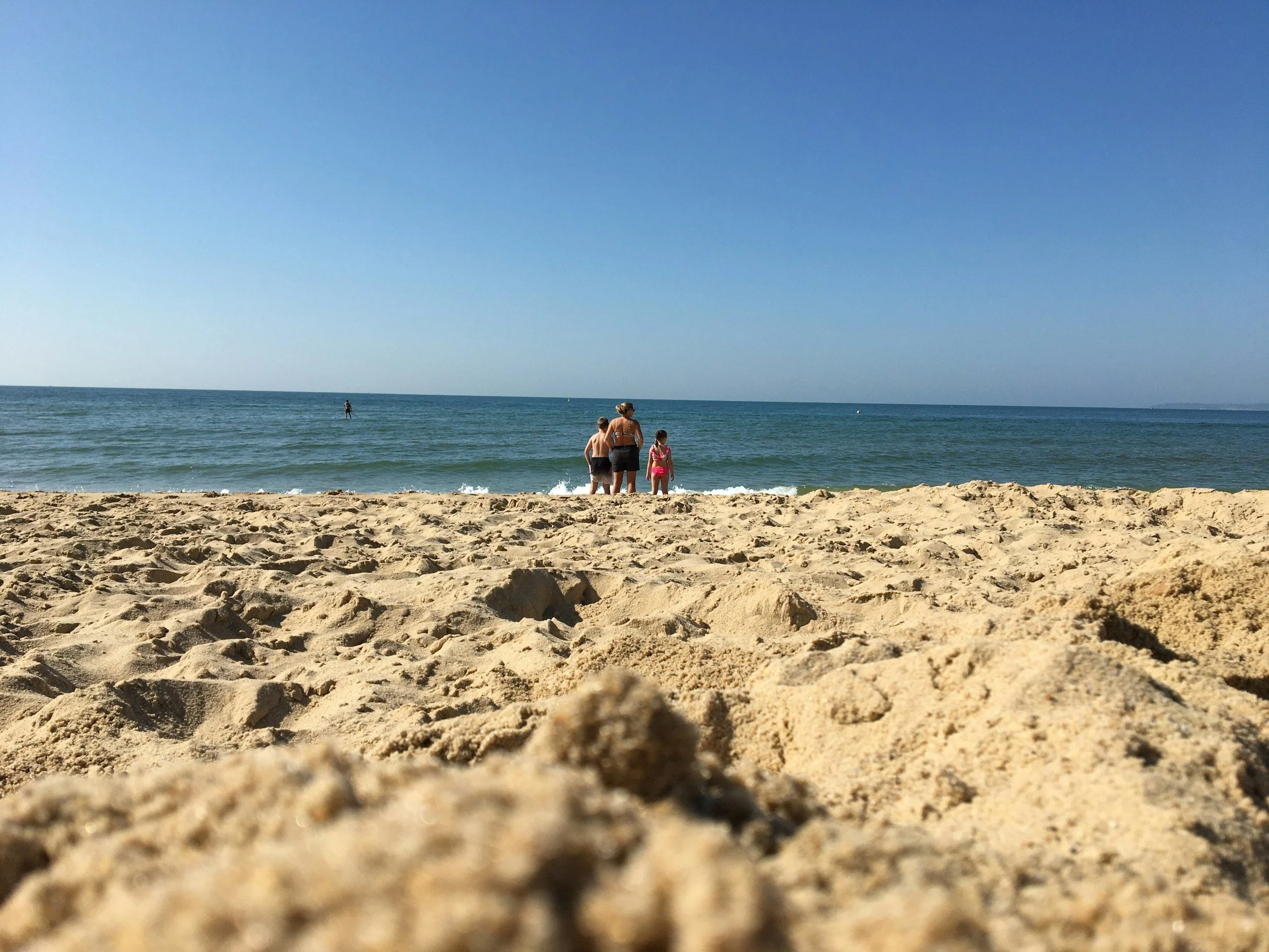 People standing on a sandy beach near the ocean, with one person paddleboarding in the distance under a clear blue sky.