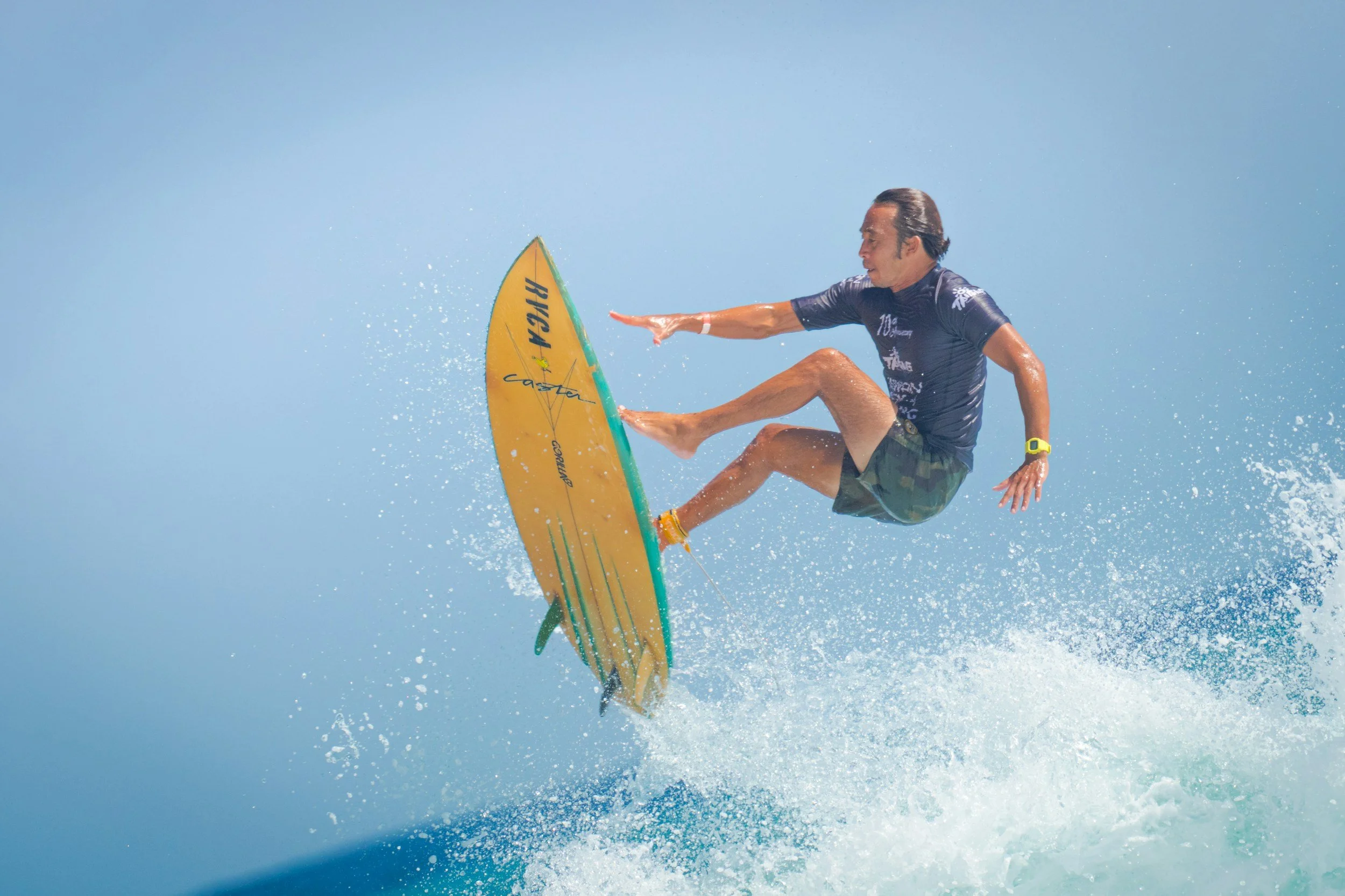 A man surfing on a yellow surfboard in the ocean, mid-air above water with splashes around, reaching out with his right hand, wearing a dark blue shirt, camo shorts, and a yellow watch.