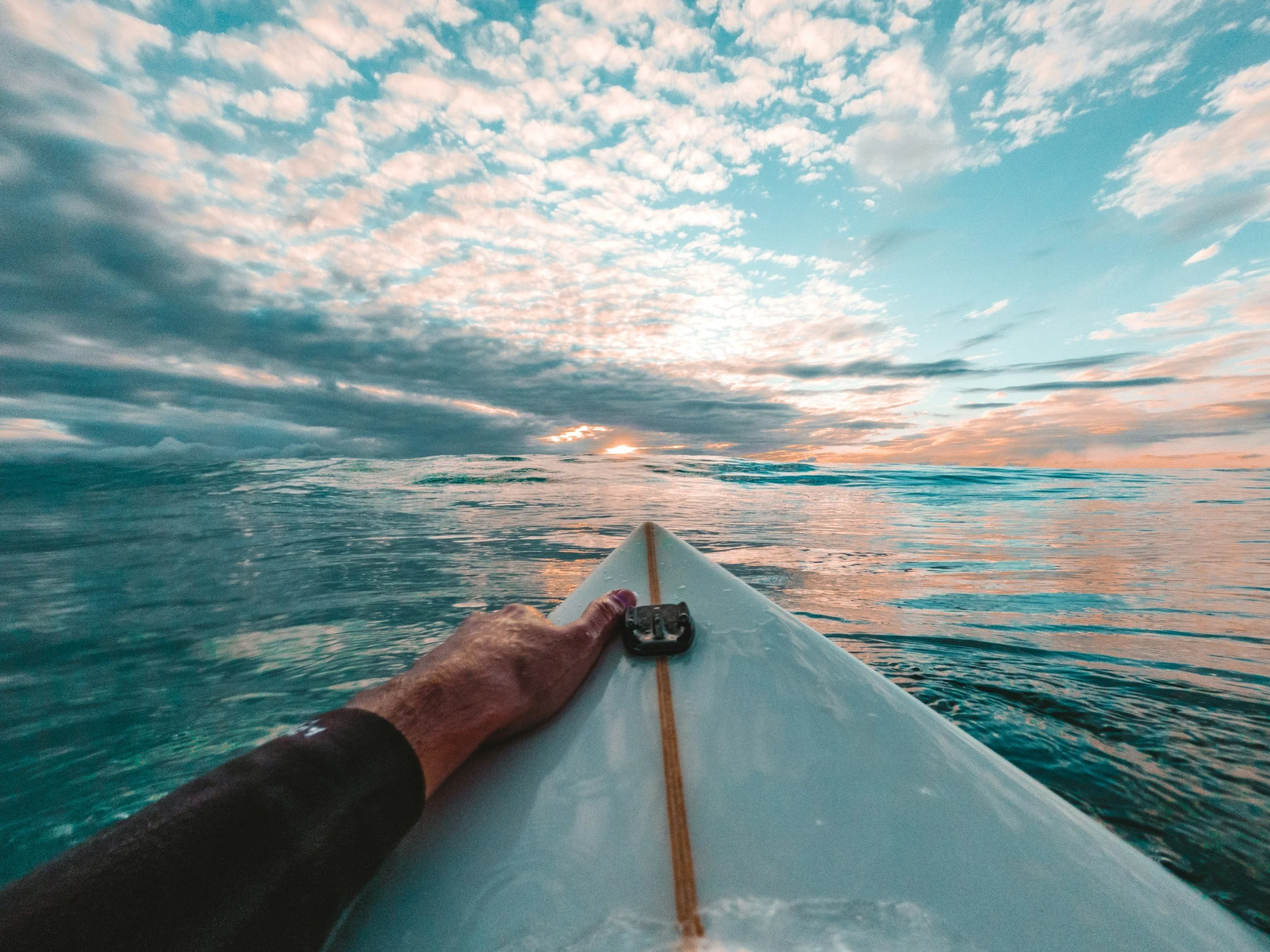 View from a surfboard on the ocean with a hand gripping the board, displaying a sunset sky with clouds.
