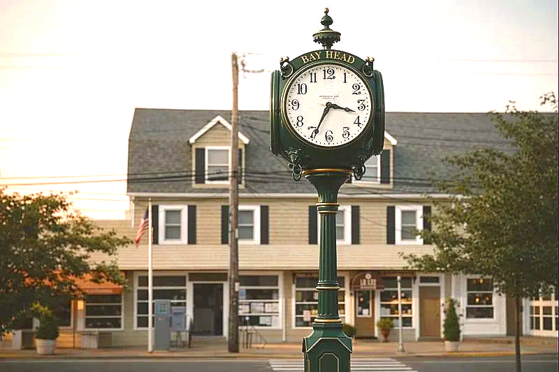 A street clock with the words 'Bay Head' on top, showing the time as 4:28, in front of a beige building with black shutters and several windows, trees, and a utility pole in the background.