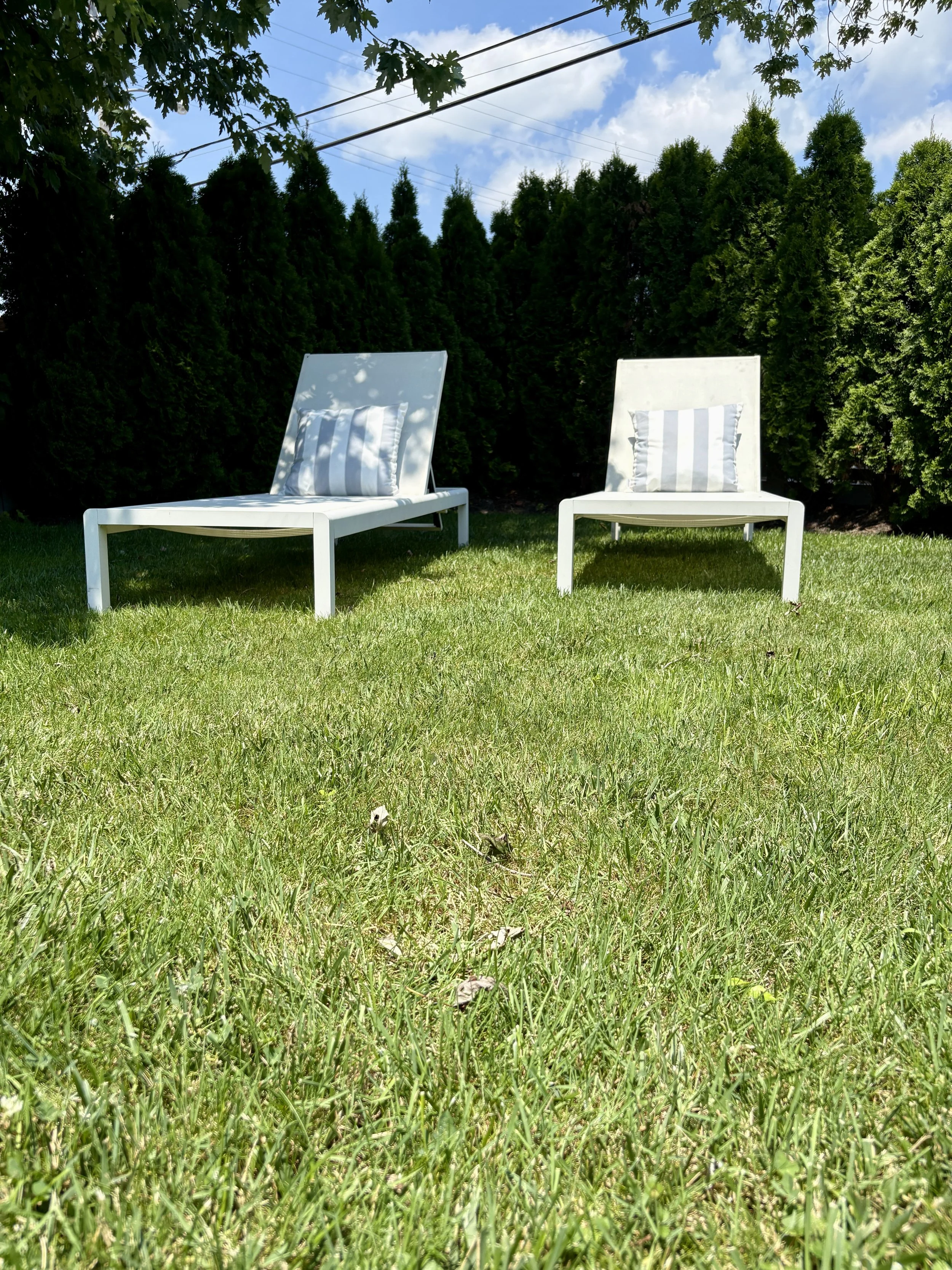 Two white lounge chairs with striped pillows on green grass in a backyard with tall trees and blue sky.