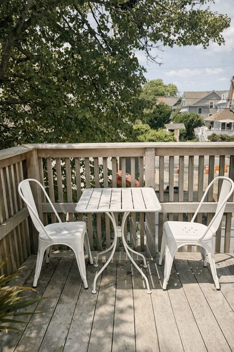 A small outdoor wooden deck with a white table and two white metal chairs, surrounded by a wooden railing and trees in the background.
