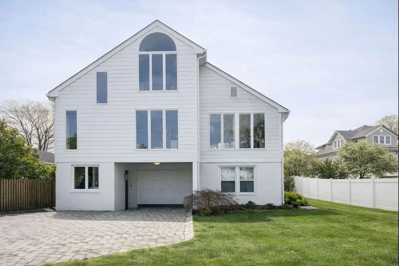 A modern white three-story house with large windows and a garage, surrounded by a green lawn and neighborhood trees.