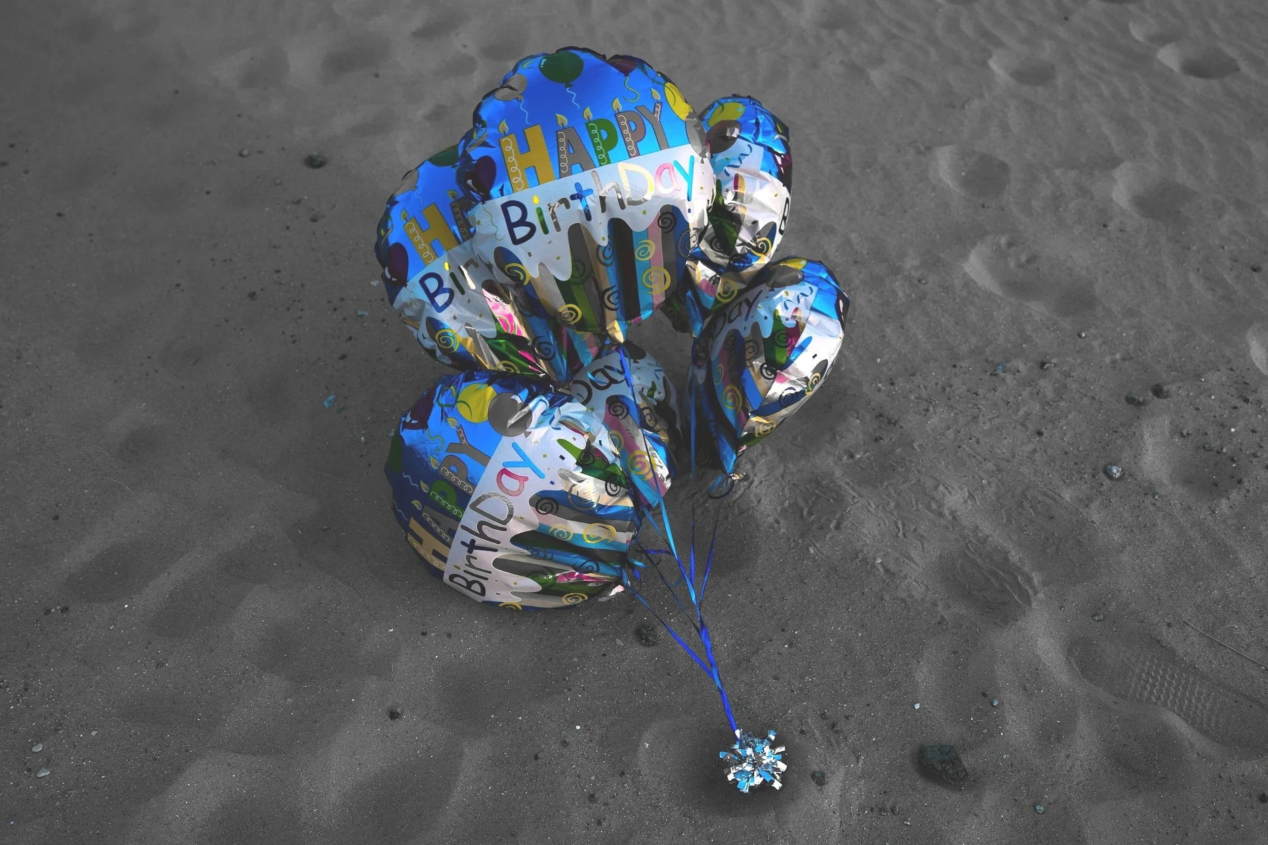 Three helium balloons with a birthday theme on a sandy surface.