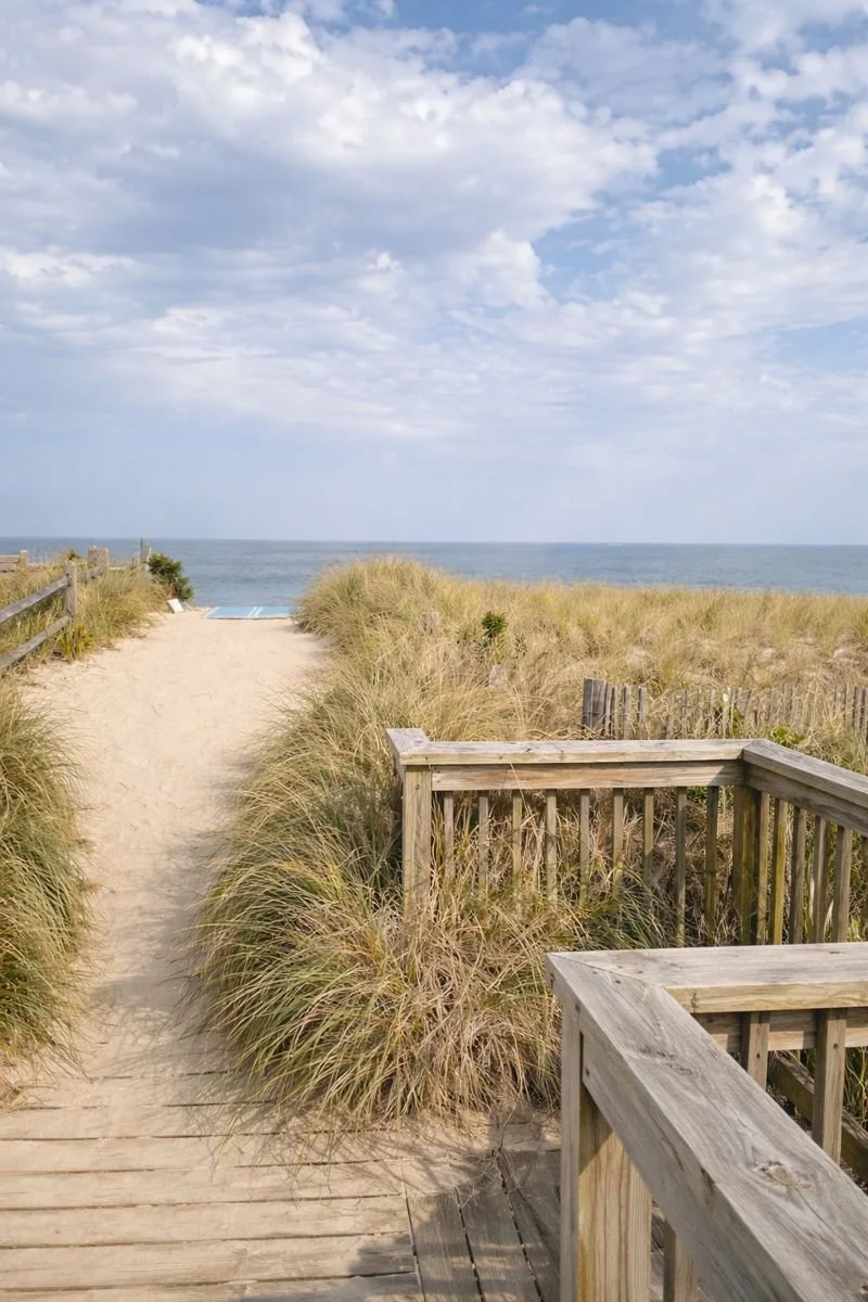 A wooden pathway with railings leading to a sandy beach with beach grass, overlooking the ocean under a partly cloudy sky.