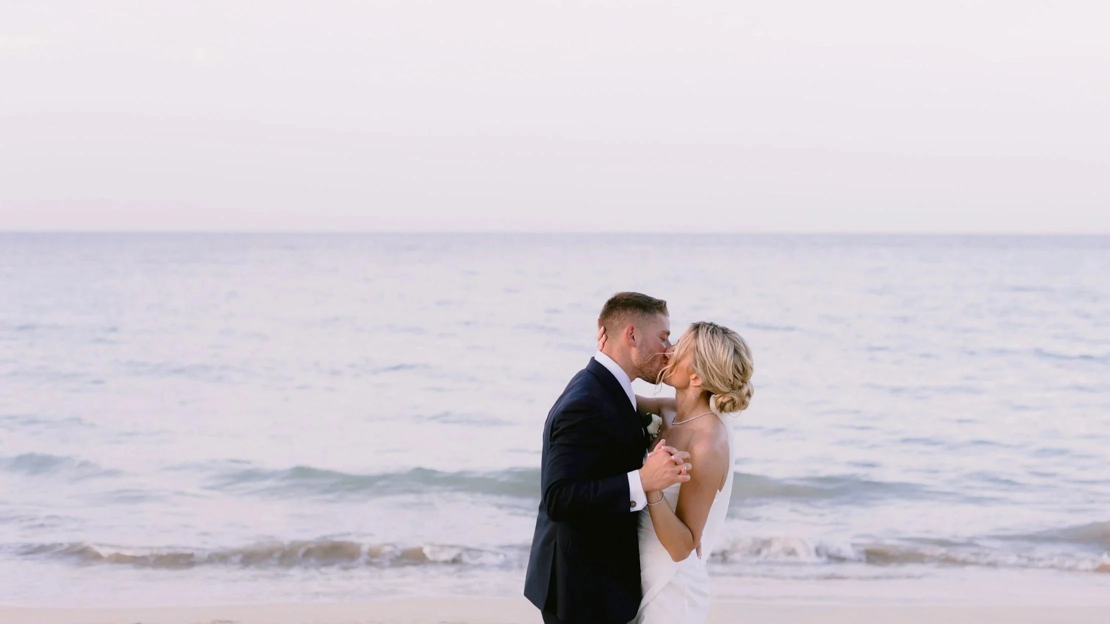 A bride and groom kissing on the beach, holding hands, with the ocean in the background.
