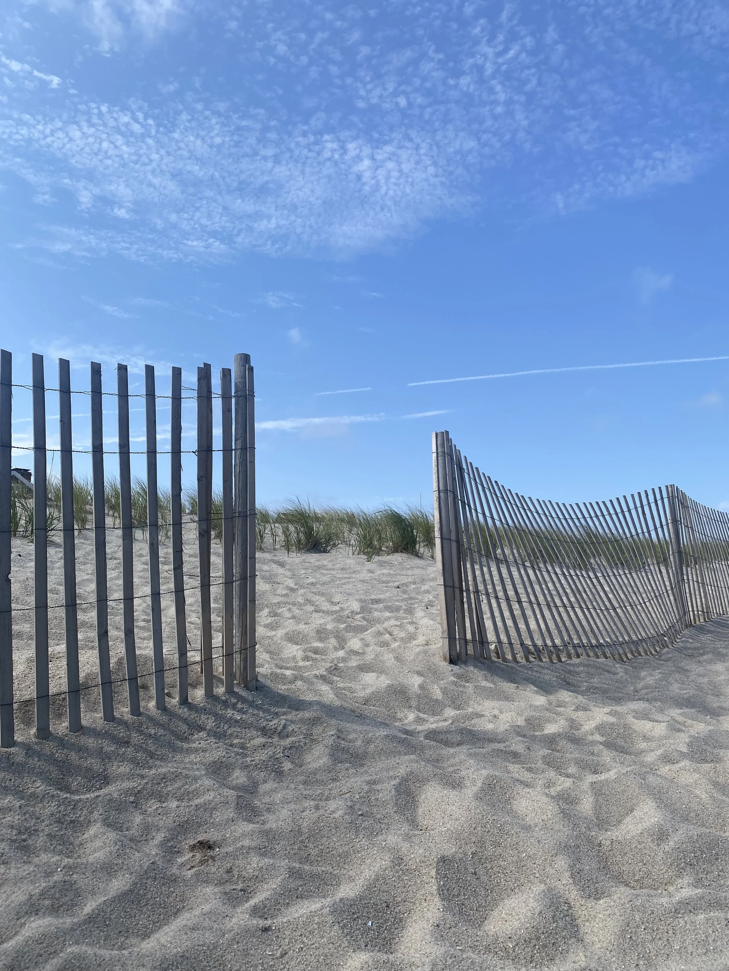 View of a sandy beach with a wooden sand fence, some green beach grass, and a partly cloudy blue sky with some wispy clouds and contrails.