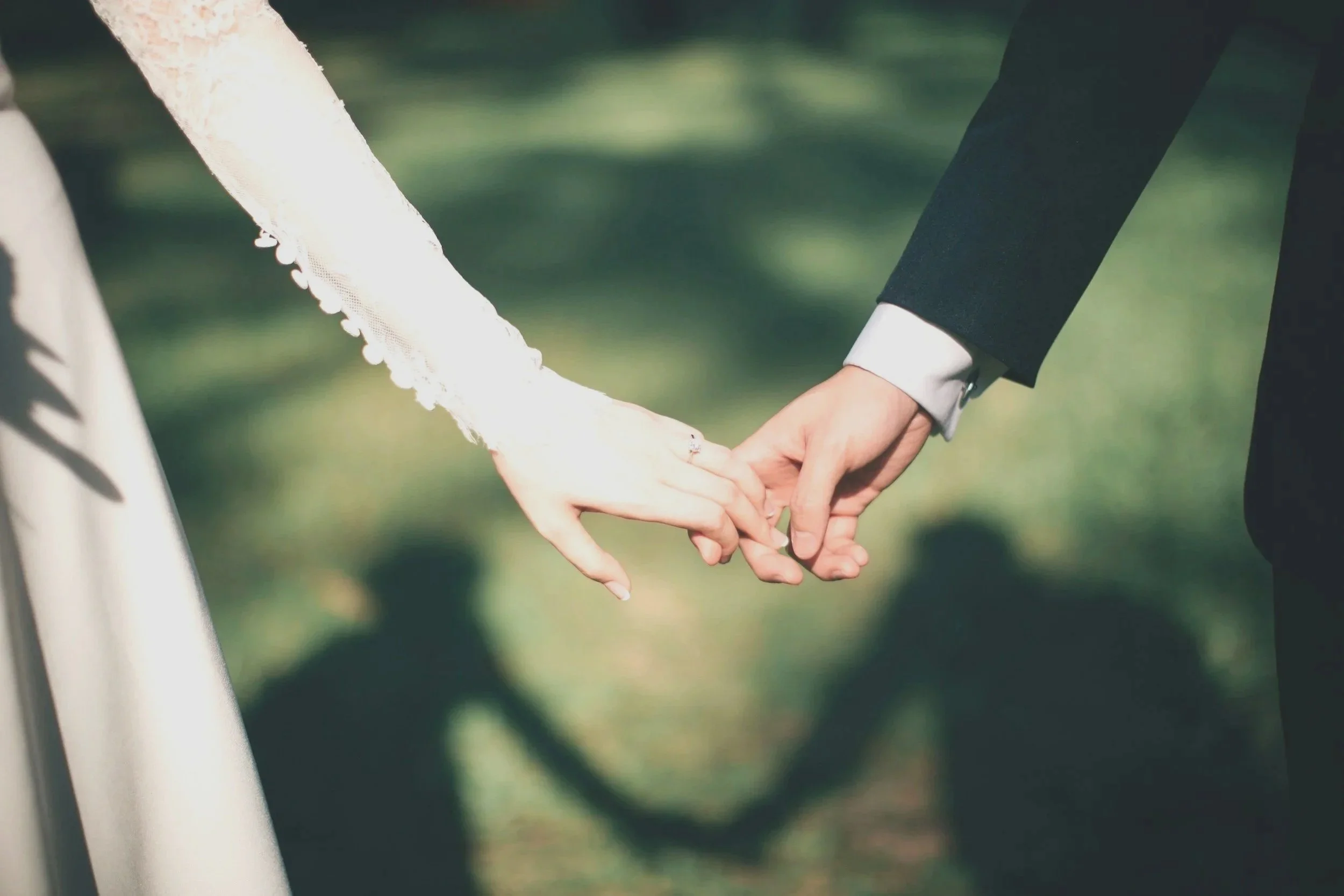 Close-up of a bride and groom holding hands outdoors, focusing on their intertwined fingers with wedding rings, in front of a blurred green background.