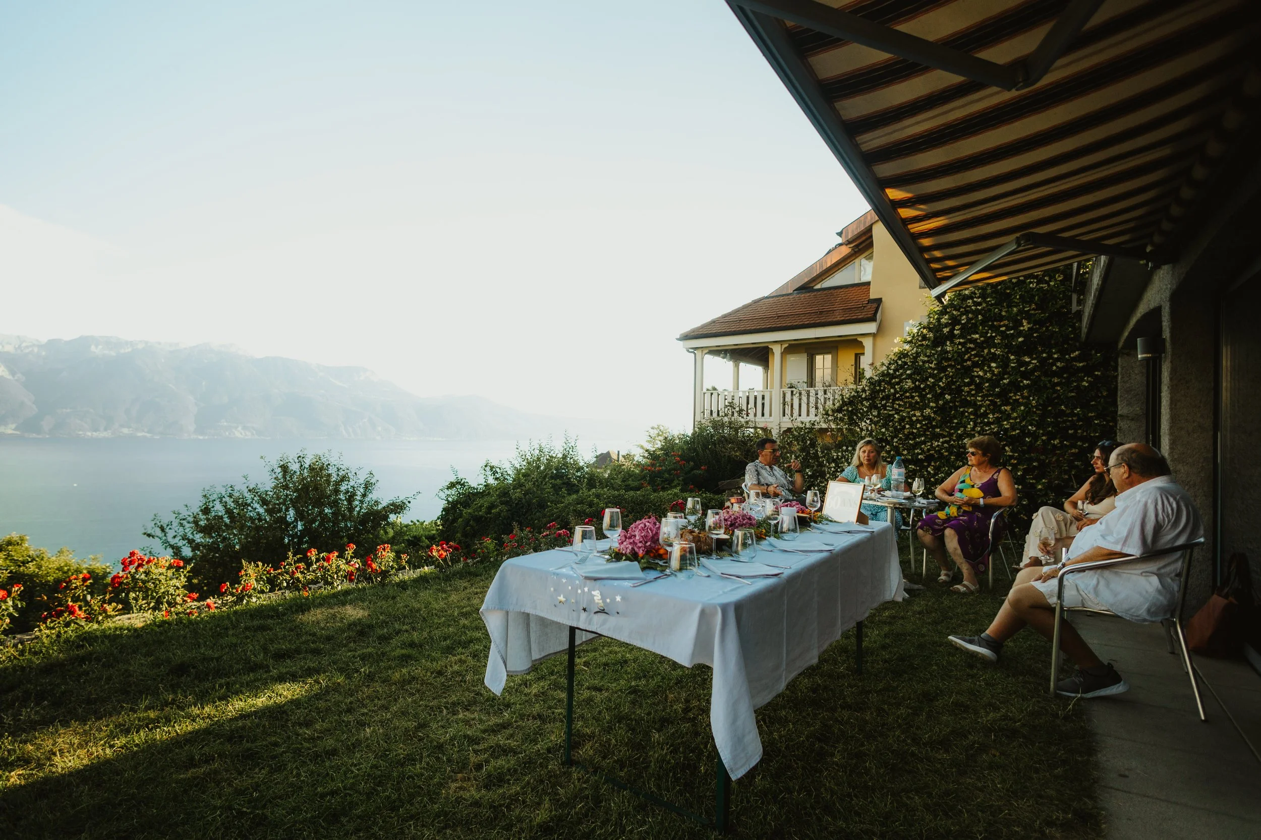 Groupe de personnes assises autour d'une table lors d'une réunion en plein air, avec vue sur un lac et des montagnes en arrière-plan, sous un auvent.
