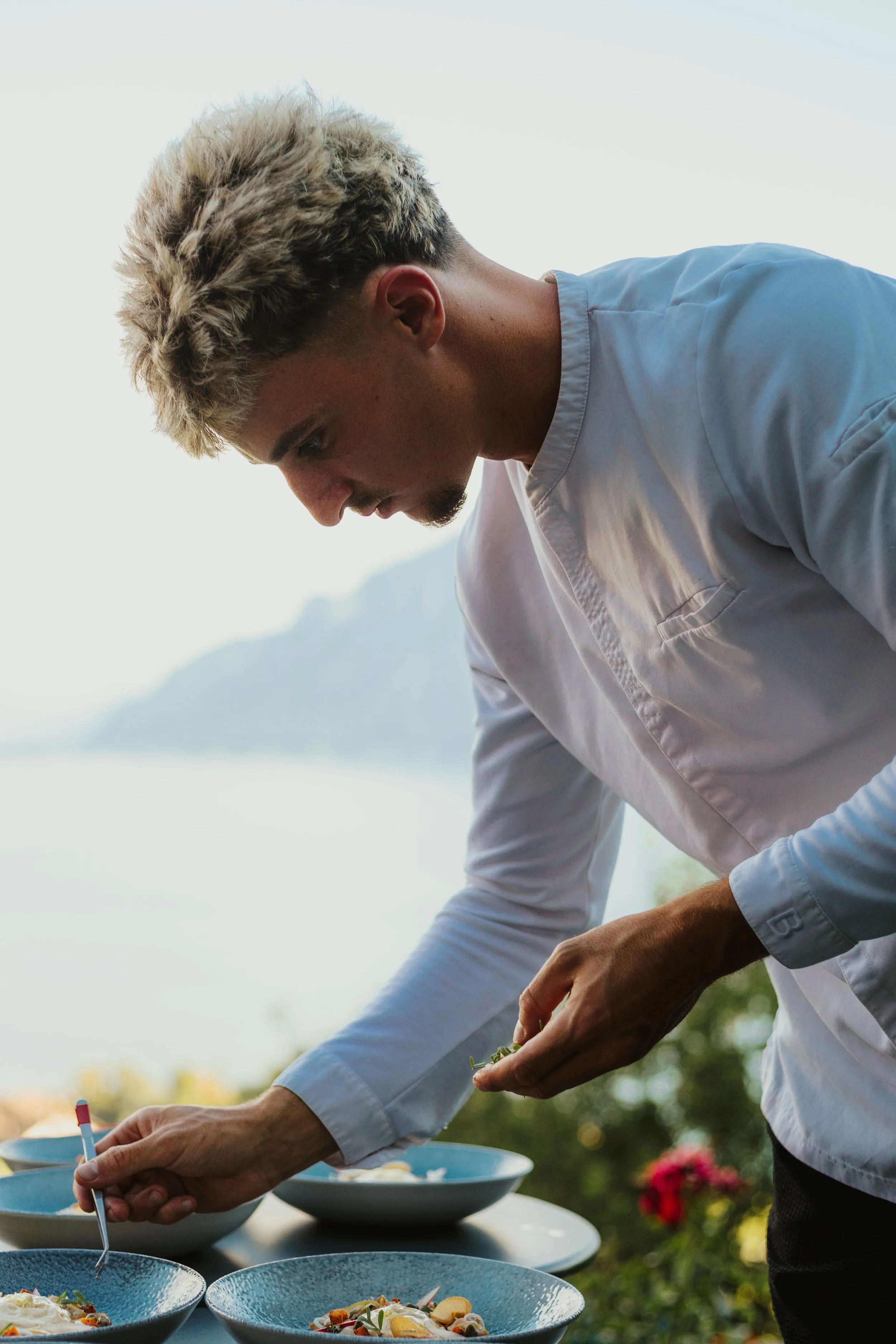 Un jeune homme en chemise blanche prépare des plats en extérieur avec une vue sur un lac et des montagnes à l'arrière-plan.