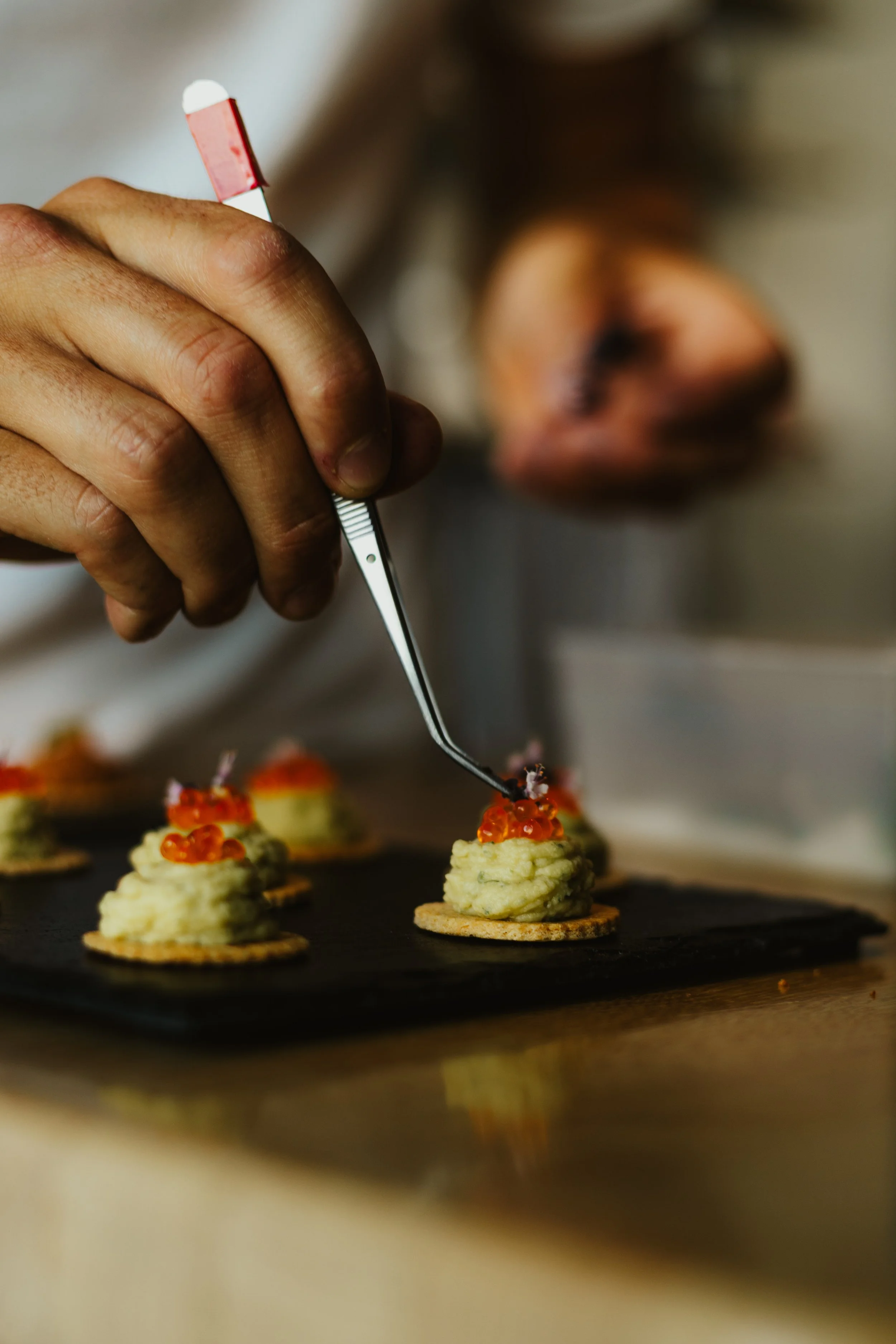 Une personne prépare des amuse-bouches avec une cuillère, en déposant des œufs de saumon sur une purée verte sur des biscuits salés.
