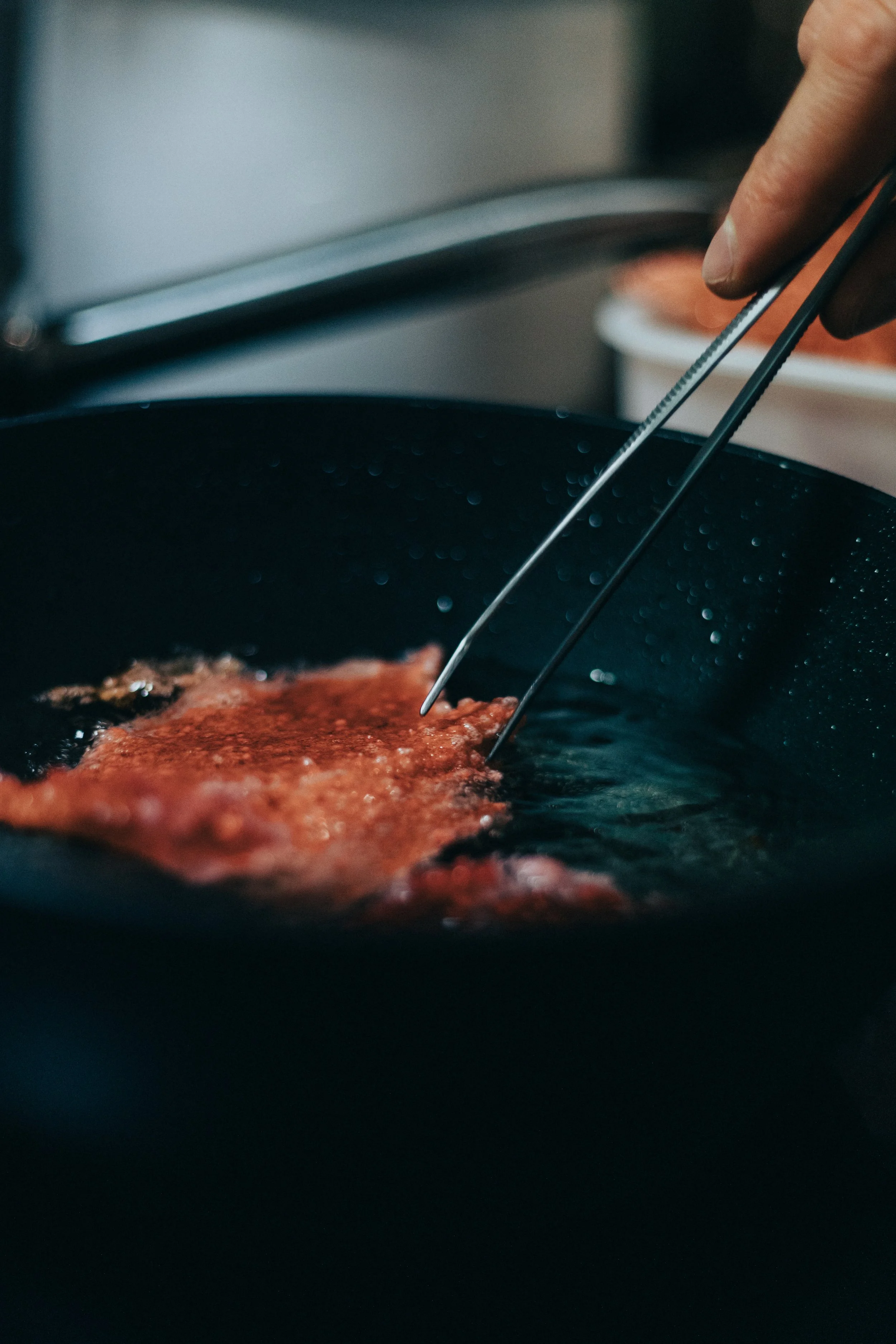 Une poêle avec de la viande en train de frire, des mains utilisant des pinces pour retourner la viande.