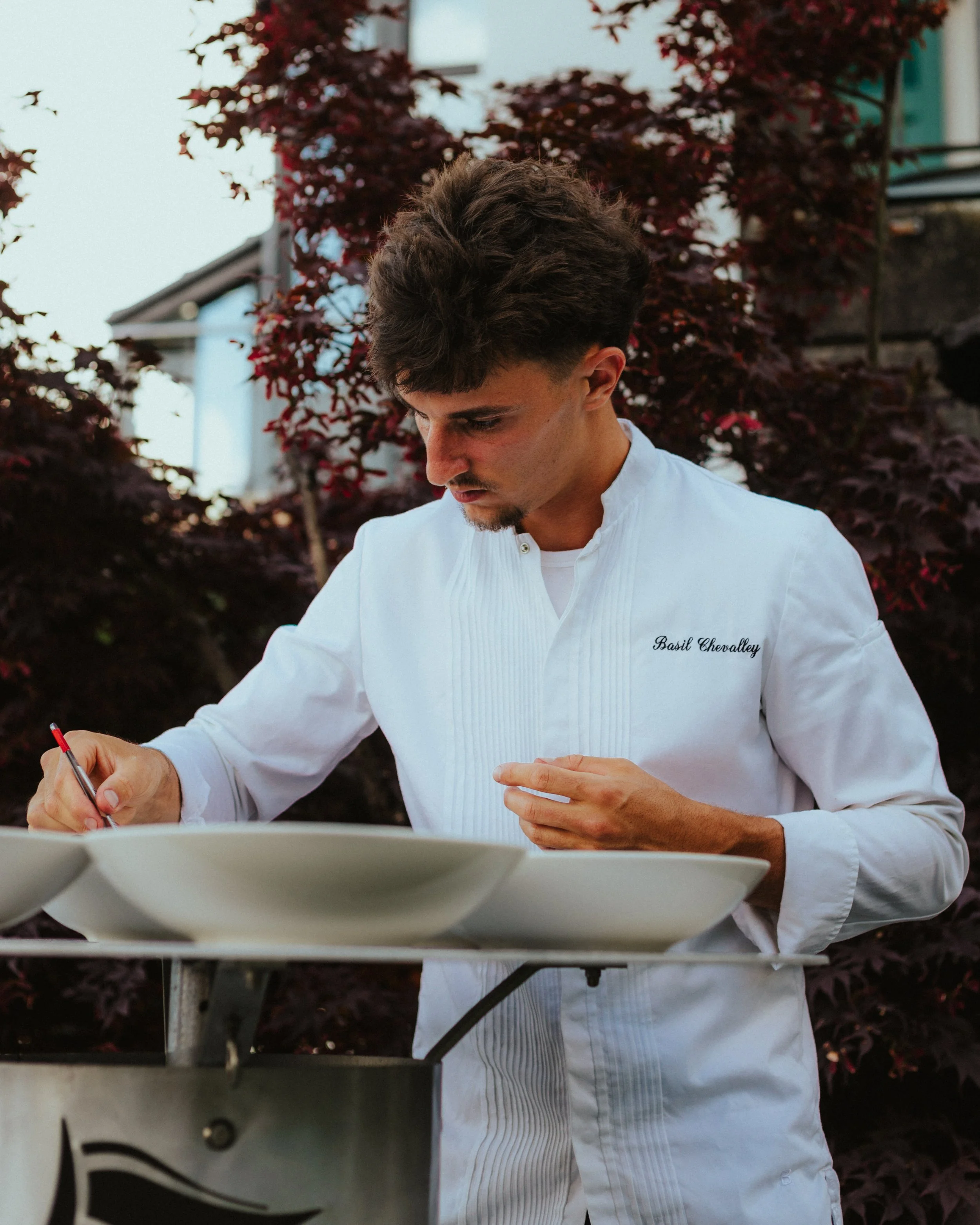Un homme habillé en chef, portant une veste blanche avec le nom 'Basil Chevalley' brodé, écrit avec un stylo sur une assiette en porcelaine, à l'extérieur près d'arbustes et d'une maison.