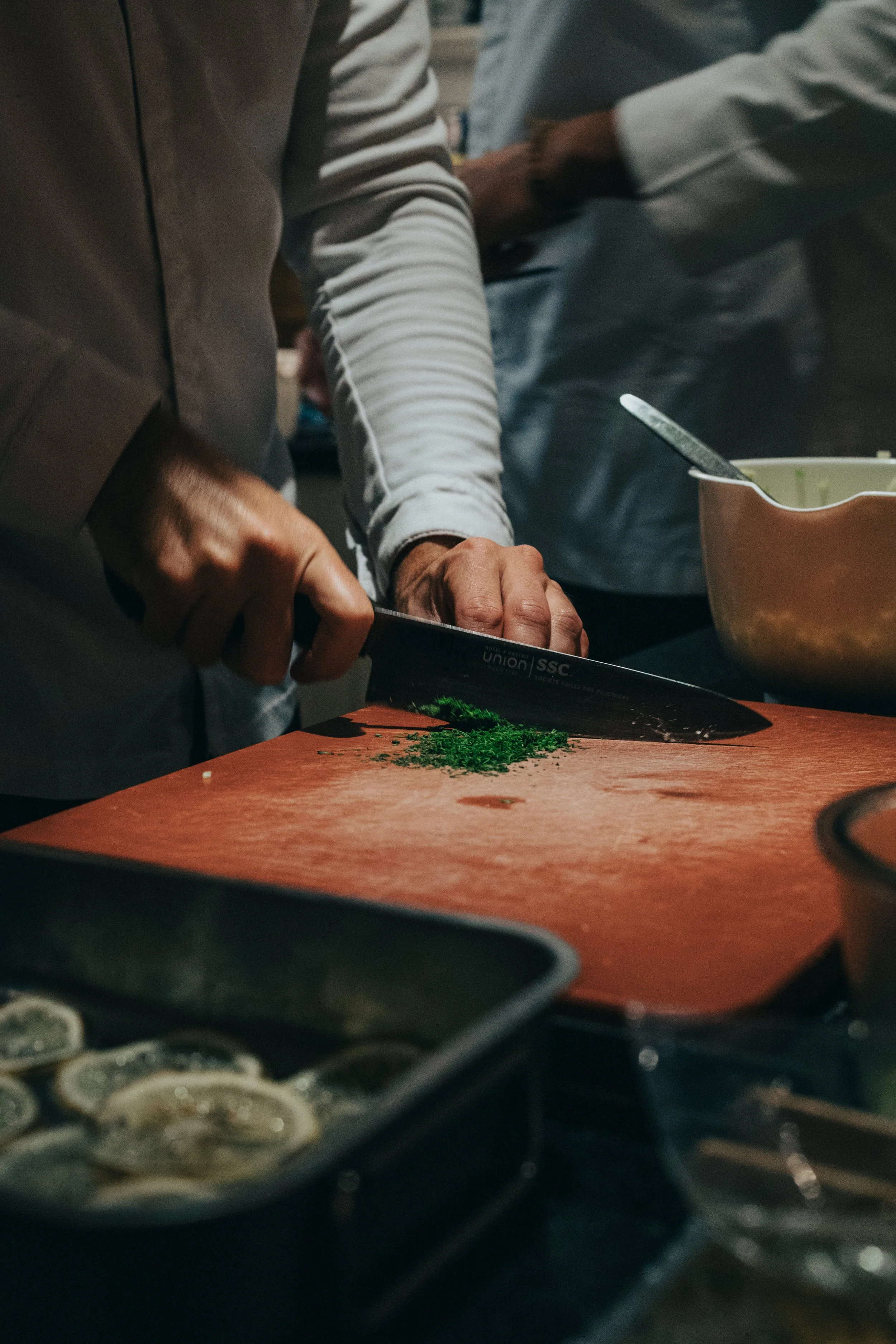 Une personne coupe des herbes vertes sur une planche en bois, dans une cuisine ou un espace de préparation alimentaire.