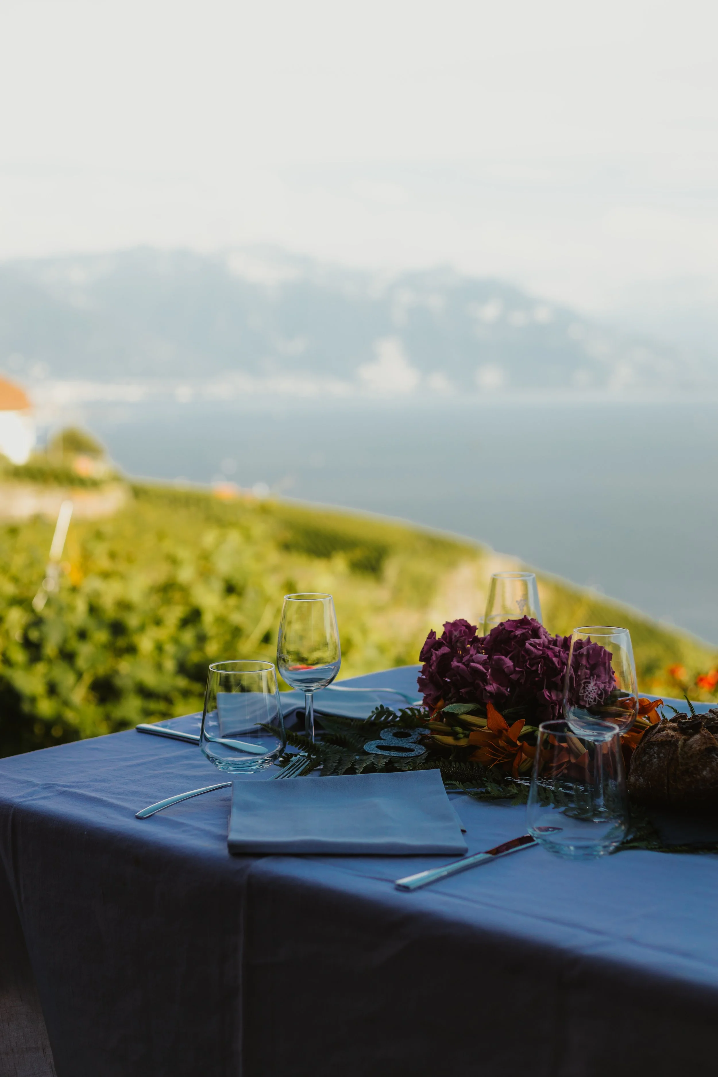 Table de dîner avec des couverts, des verres à vin, une nappe blanche, un bouquet de fleurs violettes et un pain, avec une vue panoramique sur la mer et les collines environnantes au coucher du soleil.