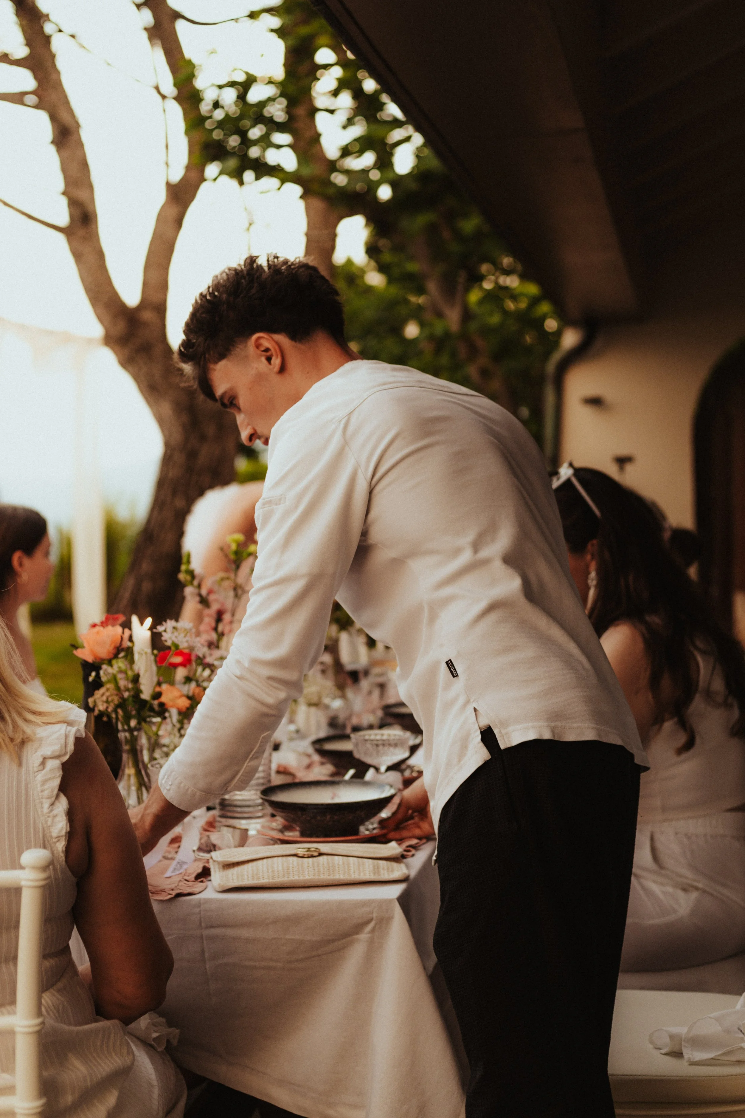 Un jeune homme en chemise blanche sert de la nourriture à une femme assise à une table de fête en extérieur, décorée de fleurs et de vaisselle élégante.