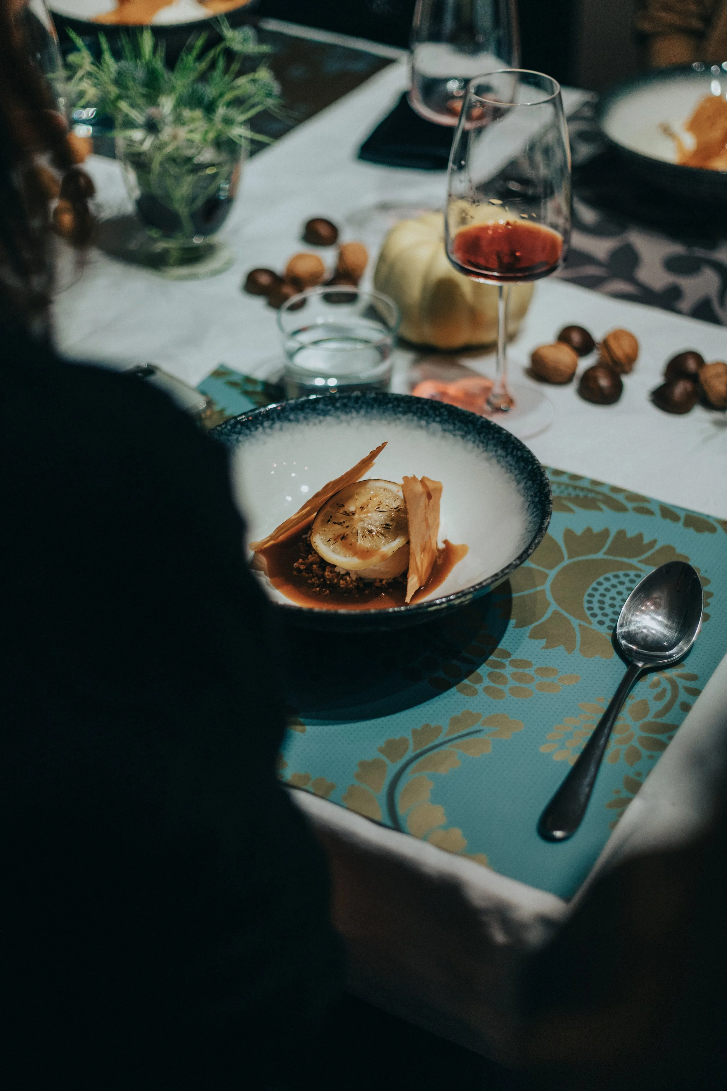 Assiette de dessert avec une rondelle de citron, deux biscuits croustillants et une sauce. Verre de vin rouge, verre d'eau, pommes et noix sur la table avec une nappe blanche et un tapis beige et bleu à motif floral.