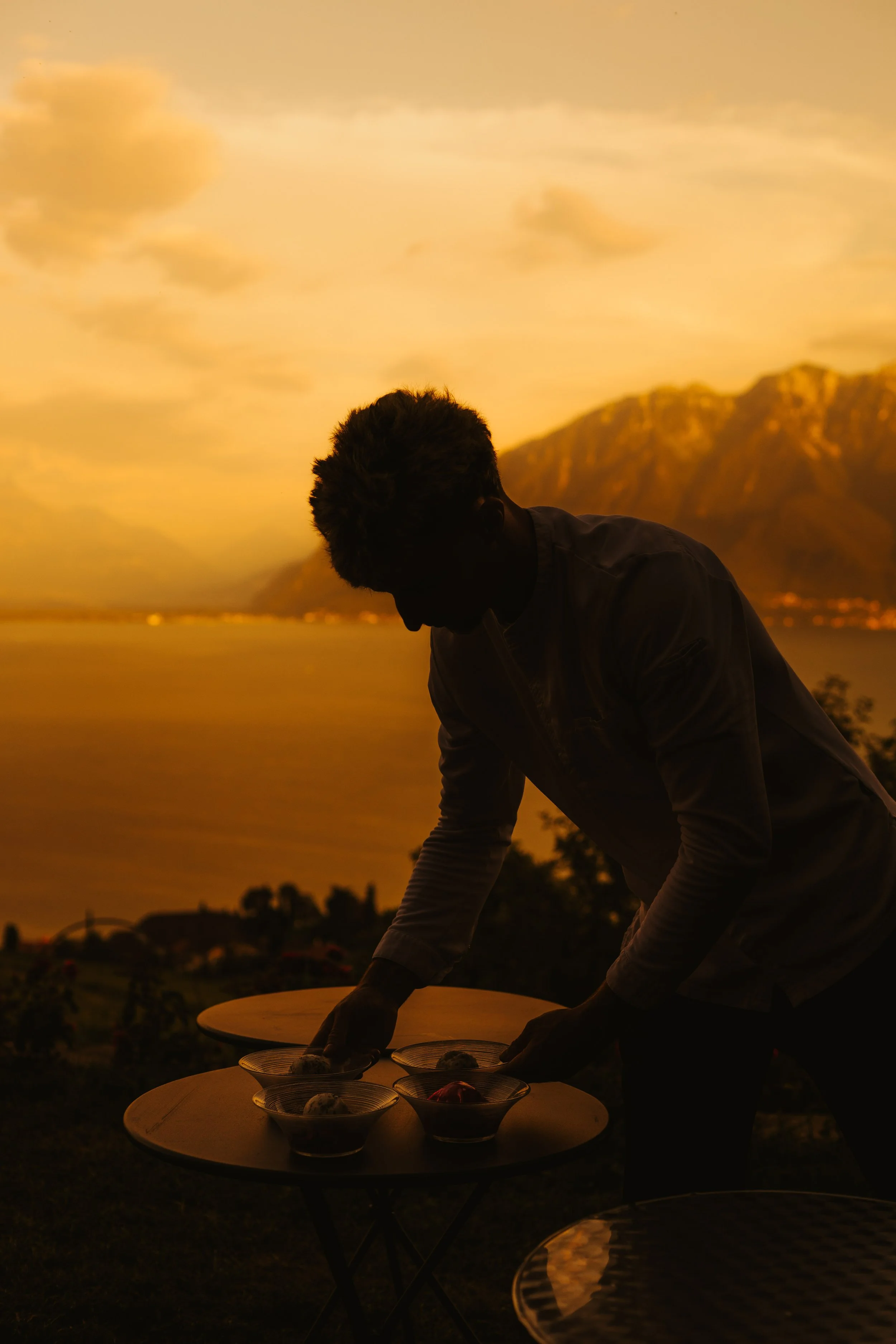 Silhouette d'un homme plaçant des bols de nourriture sur une table lors d'un coucher du soleil avec des montagnes en arrière-plan.
