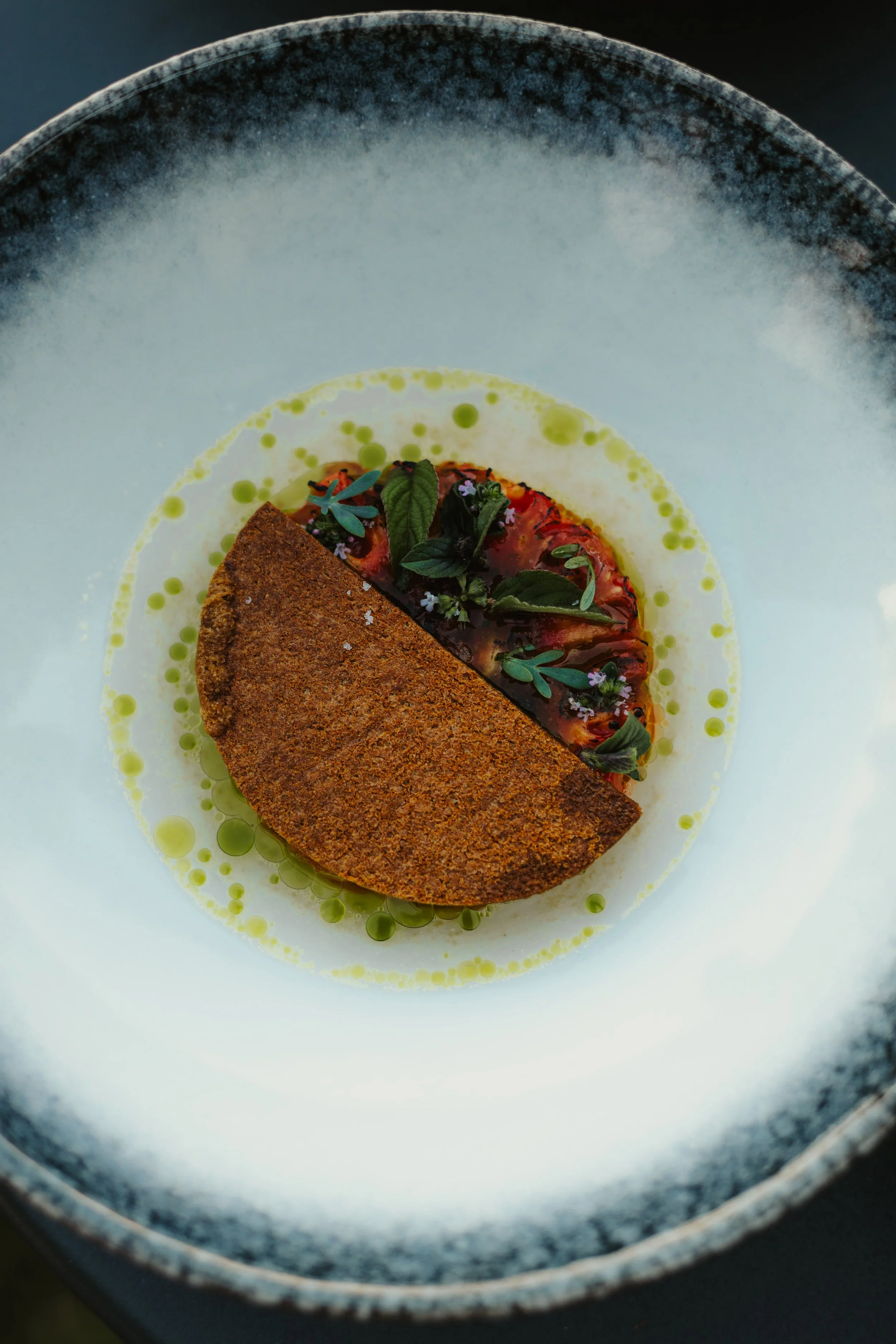 Un plat gastronomique avec un demi-cône croustillant et une garniture de légumes et herbes fraîches, présenté sur une assiette avec une sauce ou un liquide vert.