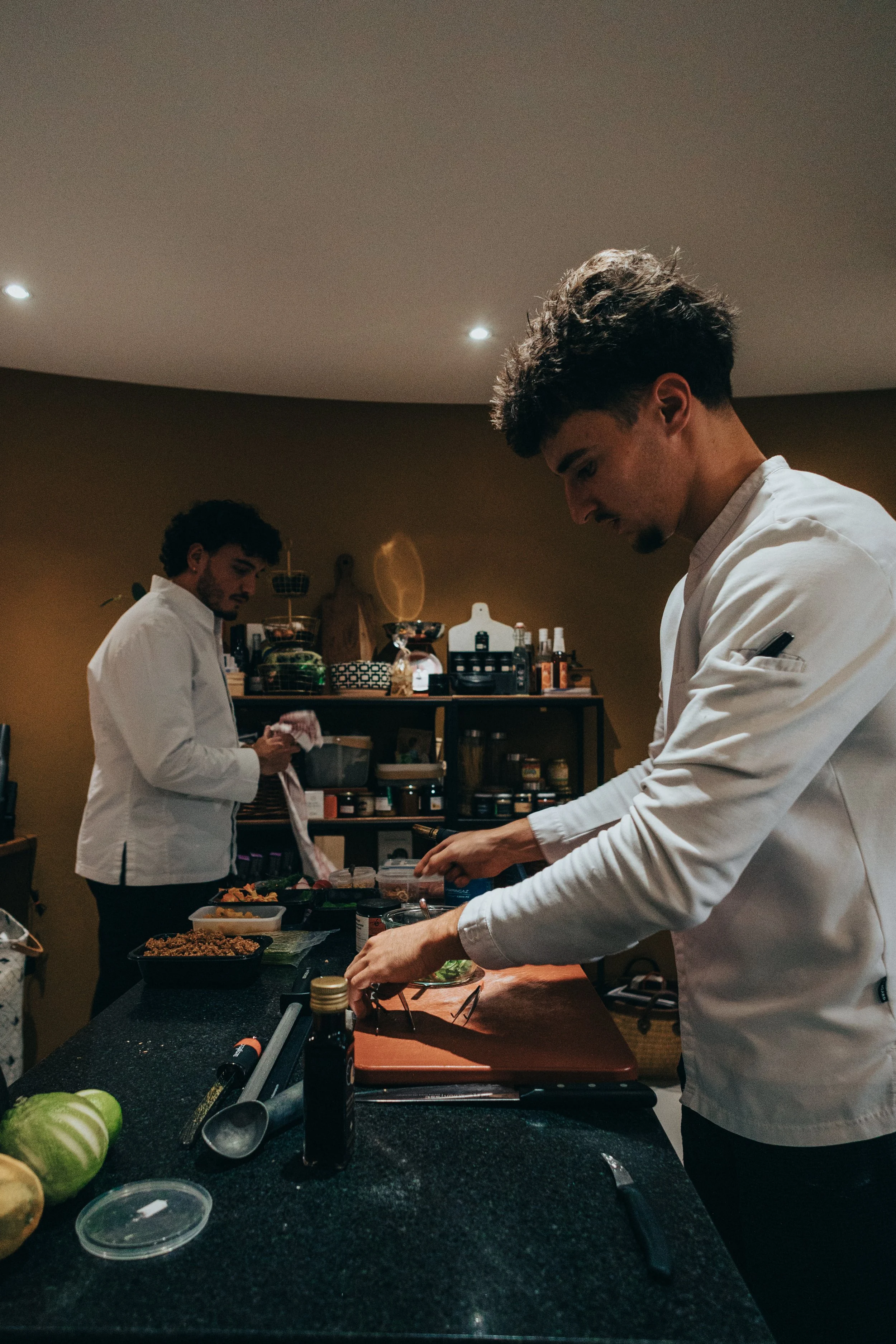 Deux jeunes hommes avec des blouses blanches préparent des aliments dans une cuisine domestique, avec des ingrédients et des ustensiles sur un comptoir.